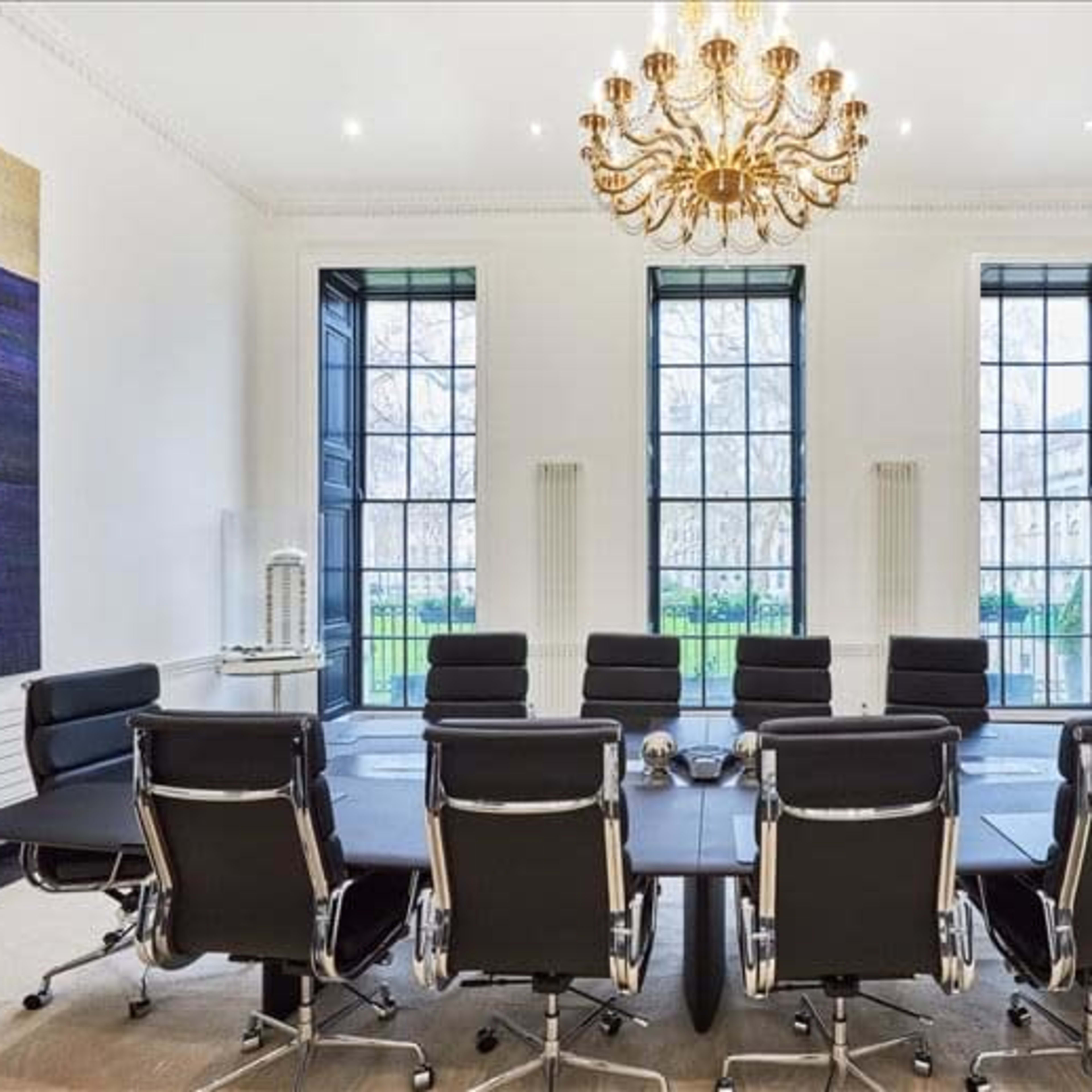 A modern conference room features a long table surrounded by black leather chairs, tall windows allowing natural light, and a large chandelier overhead.