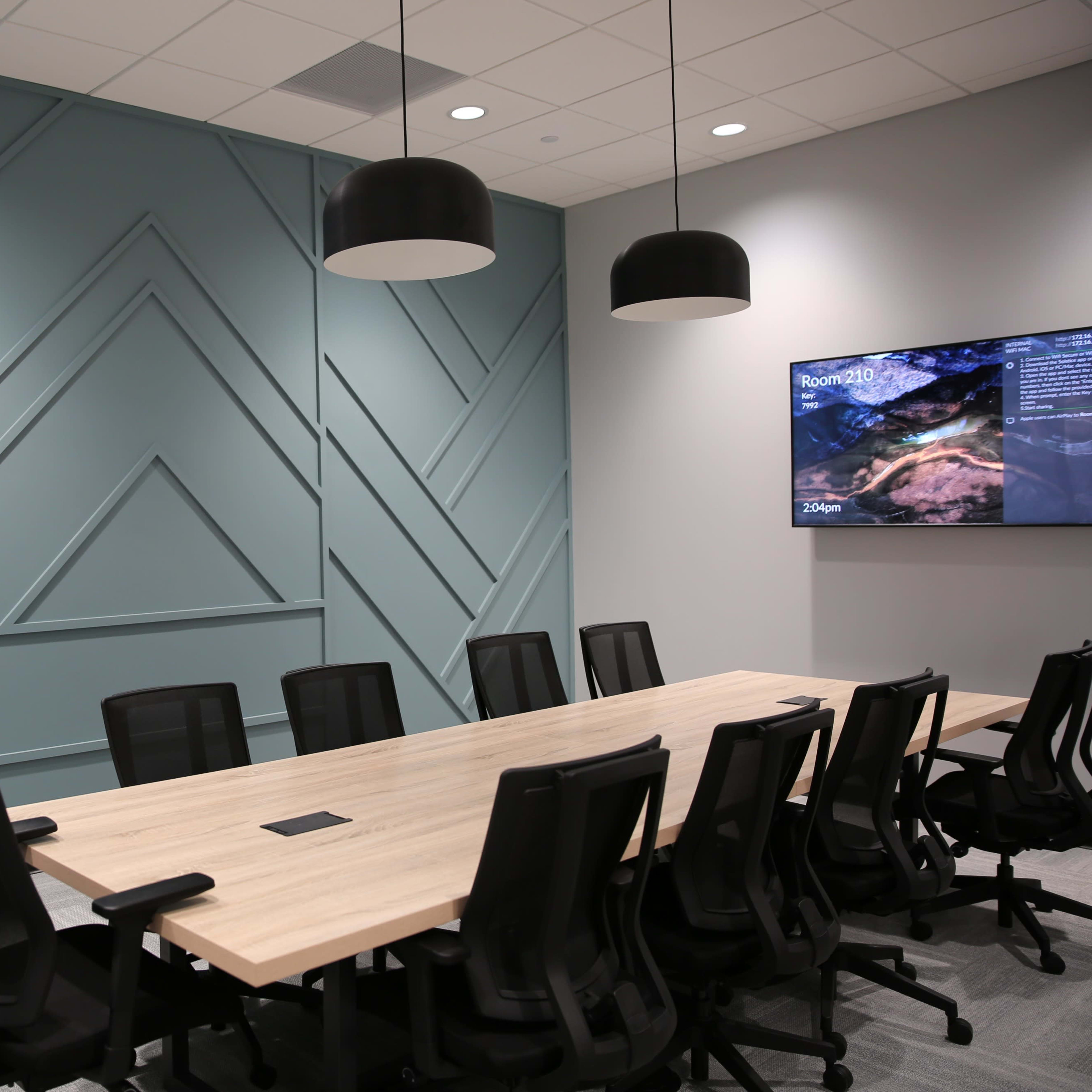A modern conference room featuring a long wooden table surrounded by black mesh chairs, with a textured blue-green wall and a television screen displaying information on one side.