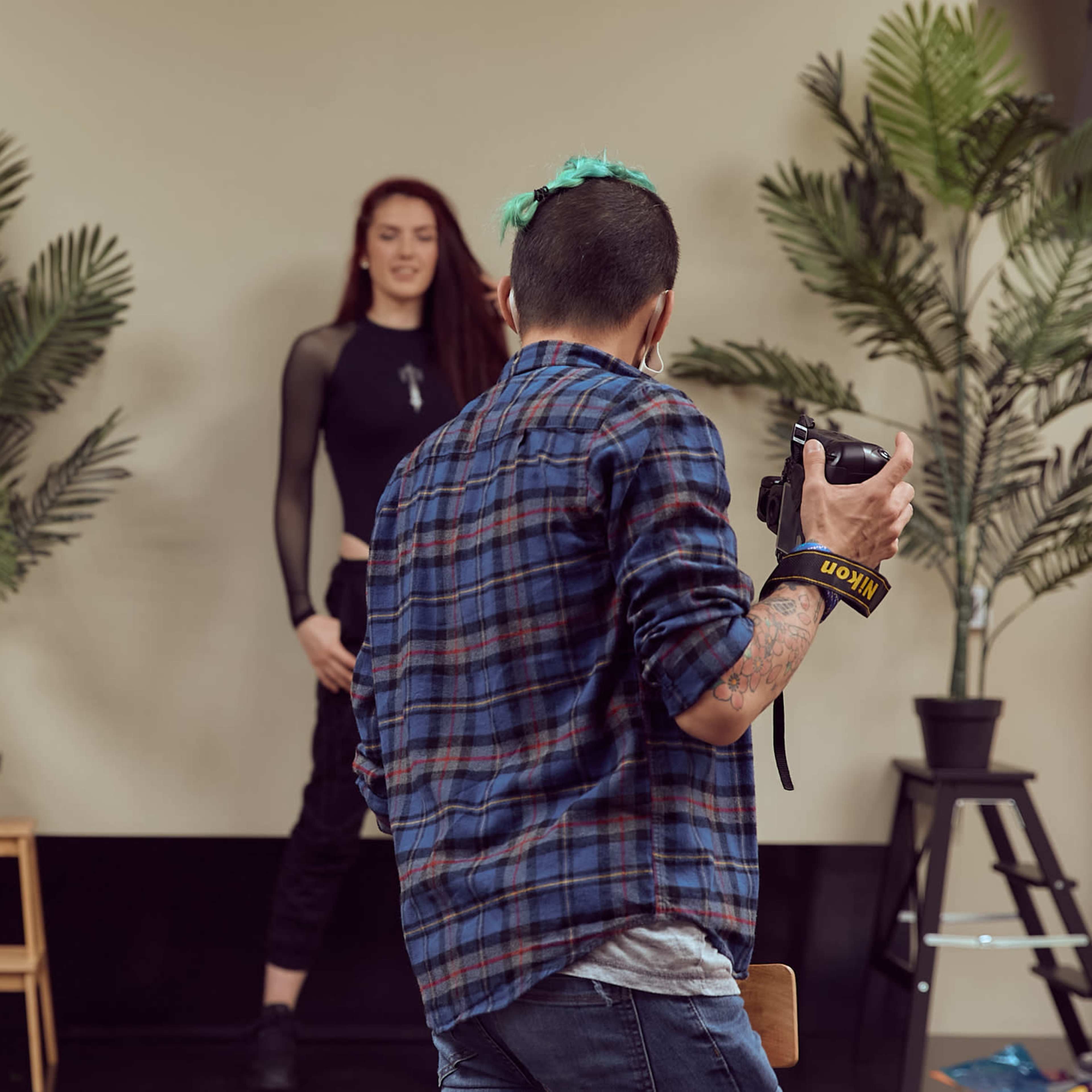A person holding a camera is photographing a model posed in front of potted plants, with a studio lighting setup in the background.