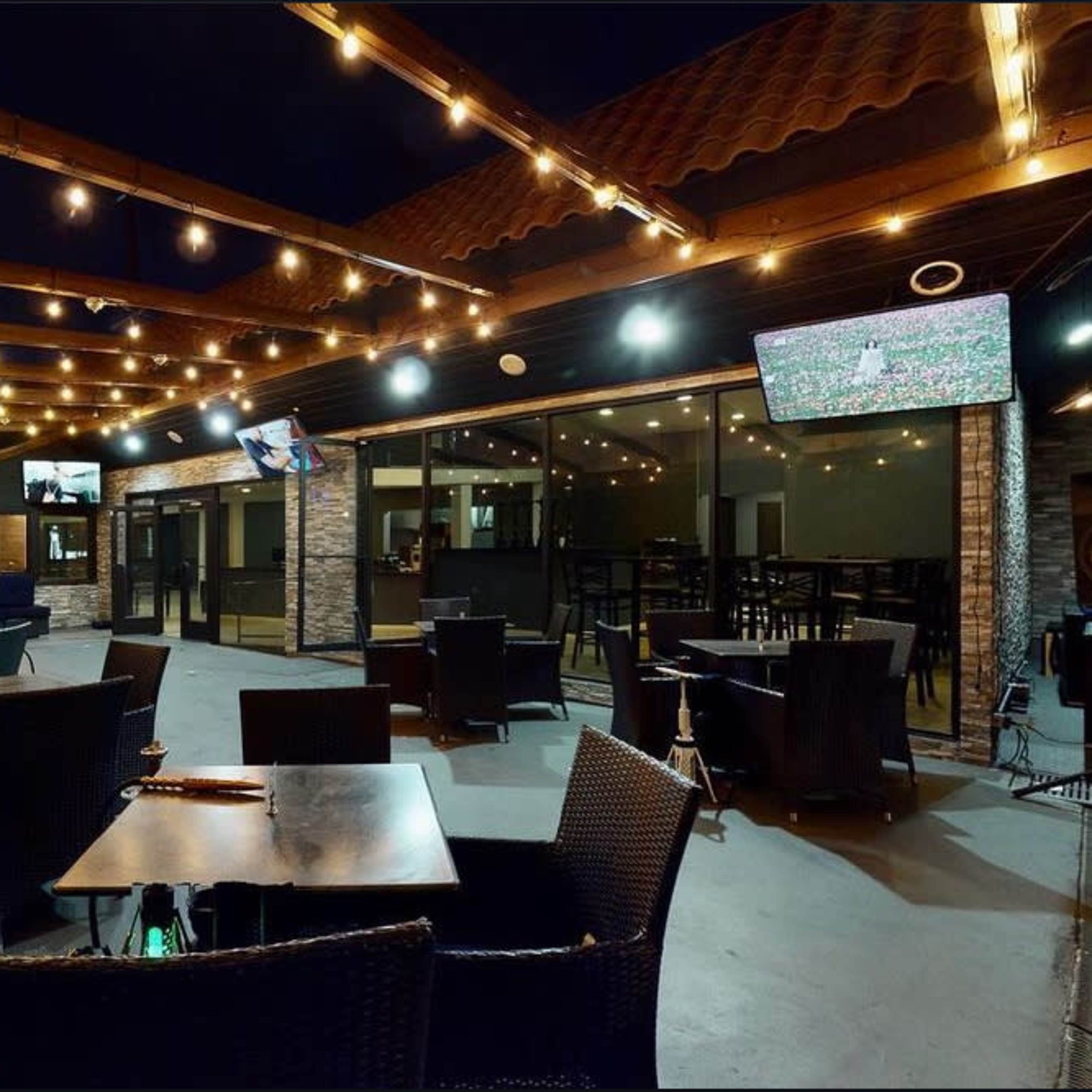 An outdoor restaurant patio with string lights, tables, and a television displaying a sports game in a nighttime setting.