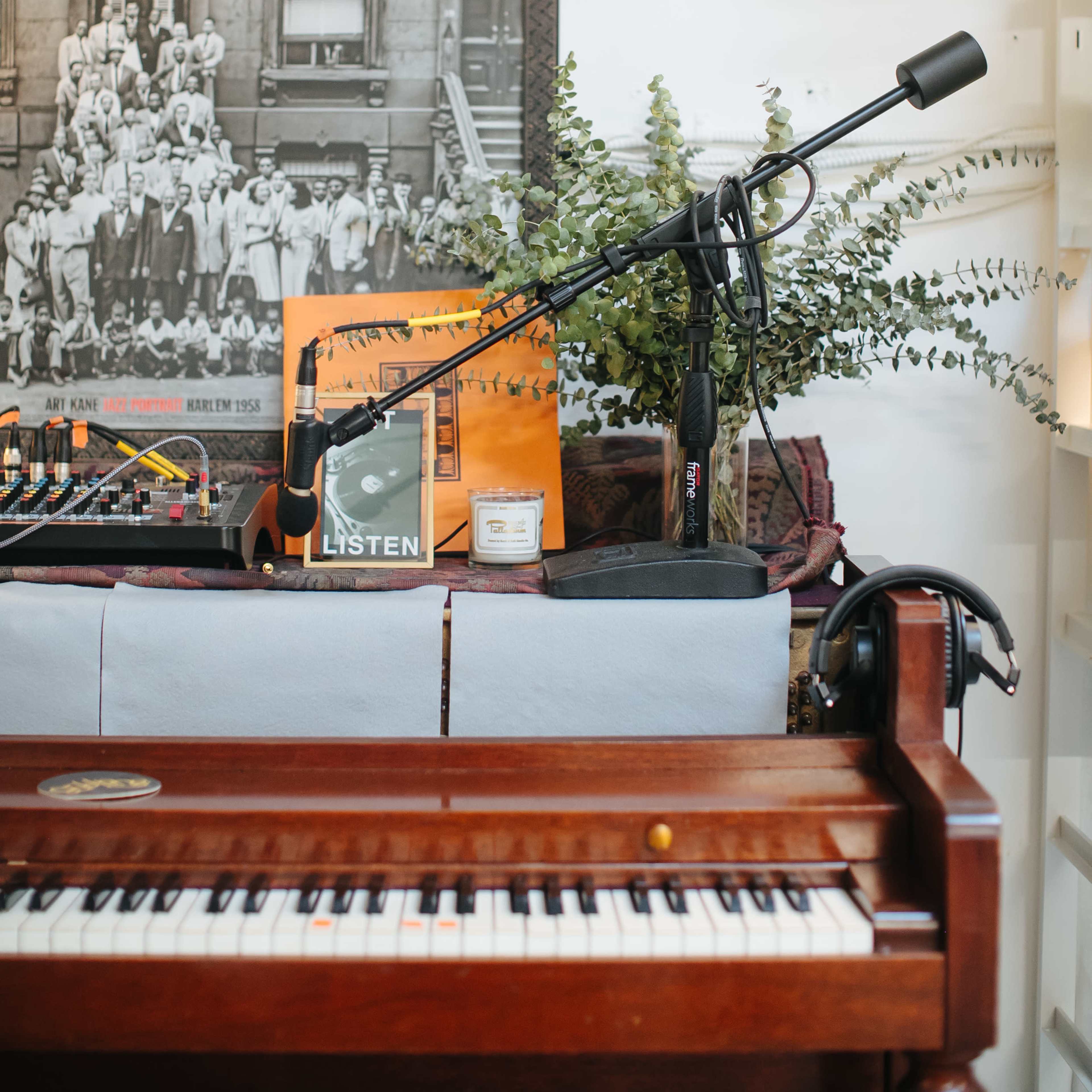 A vintage piano with a microphone and various music equipment is set against a backdrop of framed photographs and vinyl records.