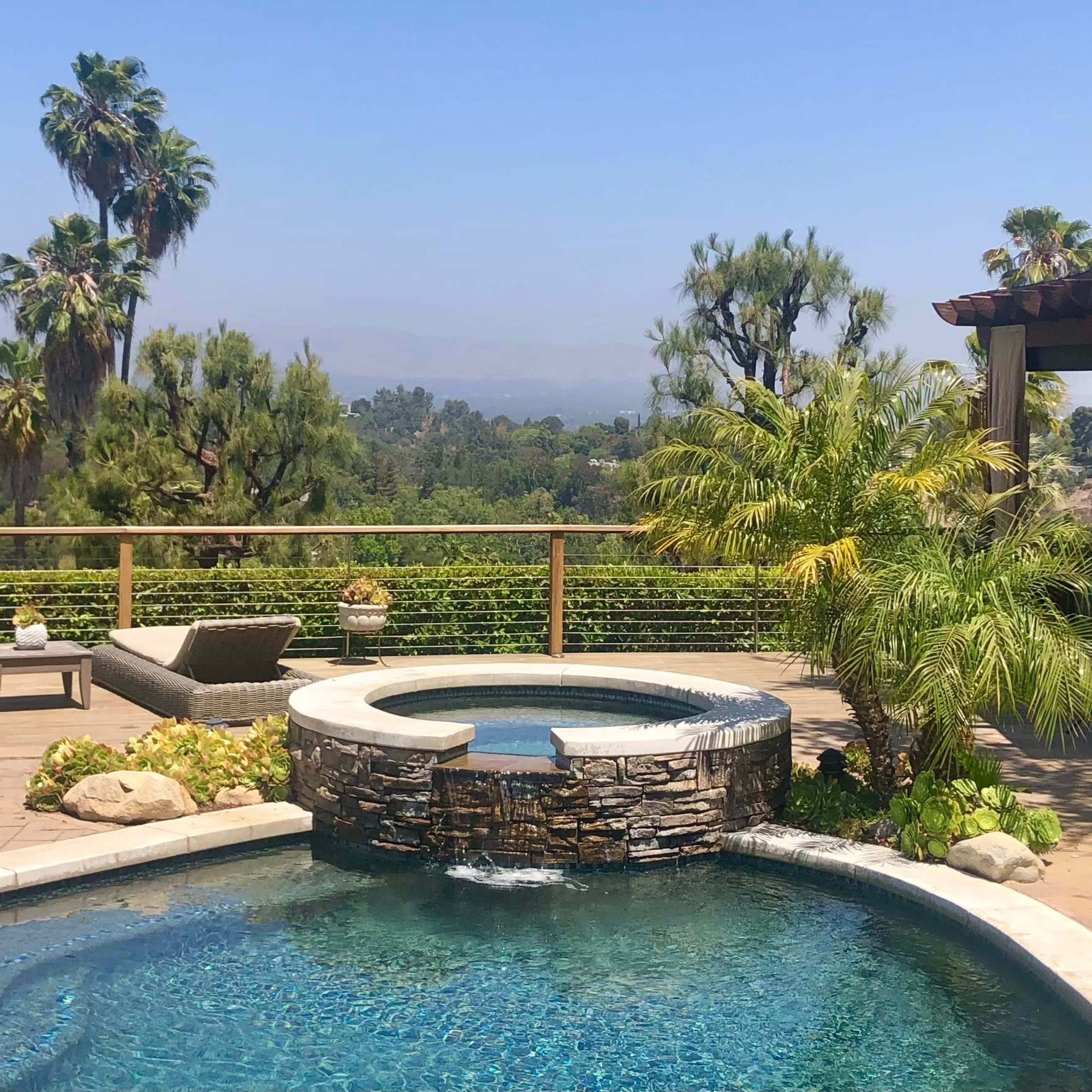 A landscaped backyard with a swimming pool featuring a stone waterfall and two lounge chairs overlooking a view of greenery and palm trees.