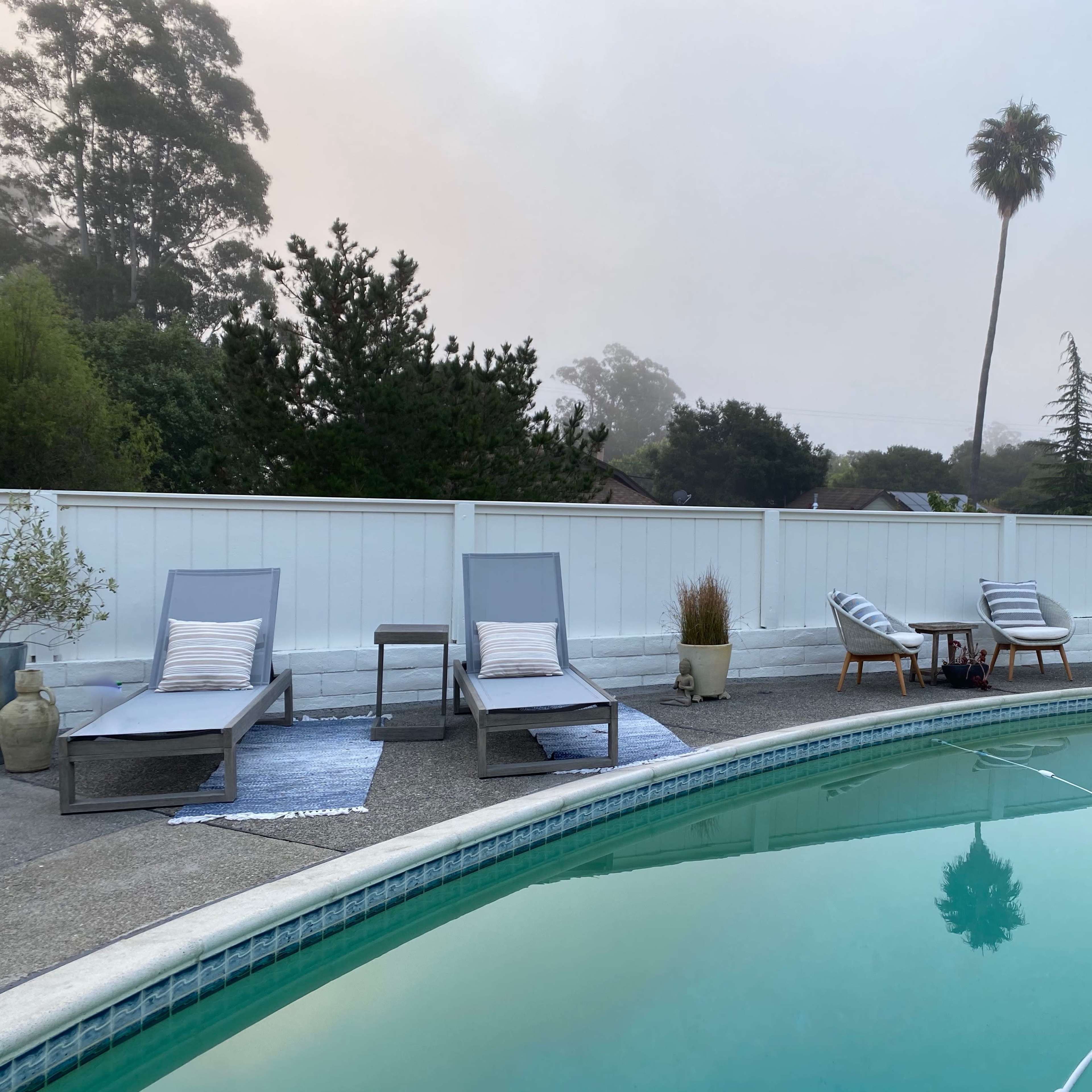 The scene features a swimming pool surrounded by lounge chairs and a small seating area, with a misty backdrop and a palm tree in the distance.