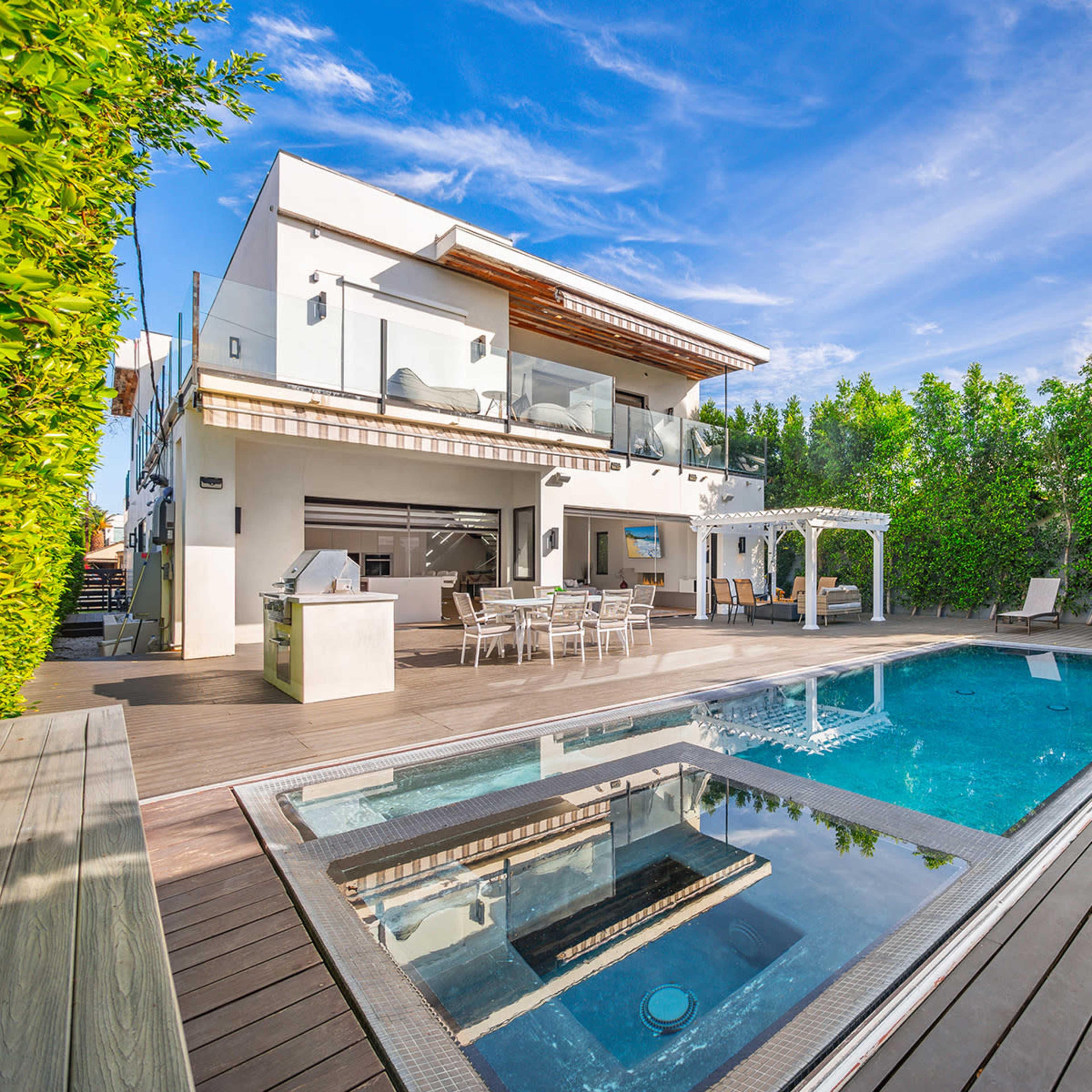 A two-story house with a modern design features a swimming pool surrounded by wooden decking and landscaped greenery.