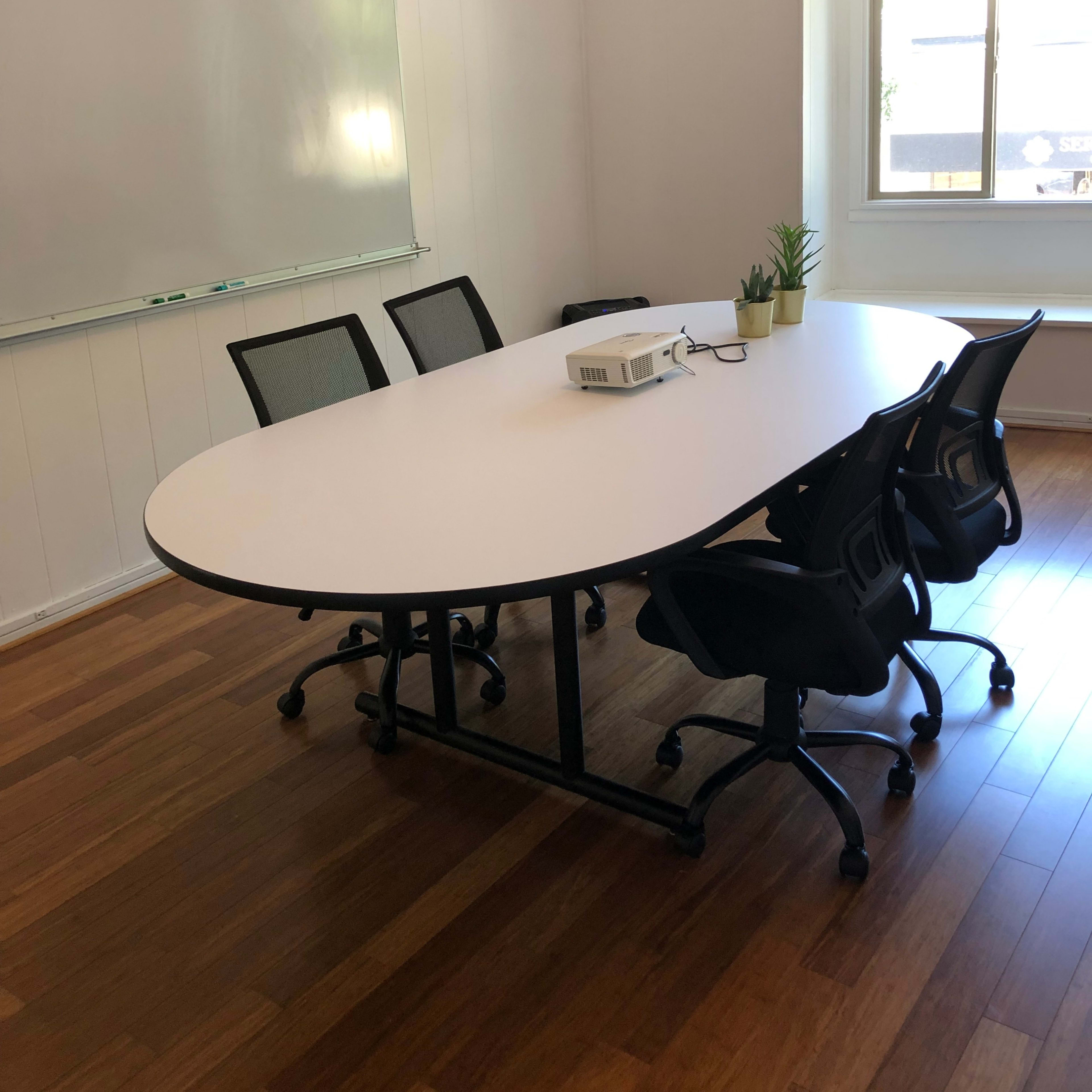 A conference room features an oval table surrounded by four chairs, a projector placed on the table, and a large whiteboard on the wall.
