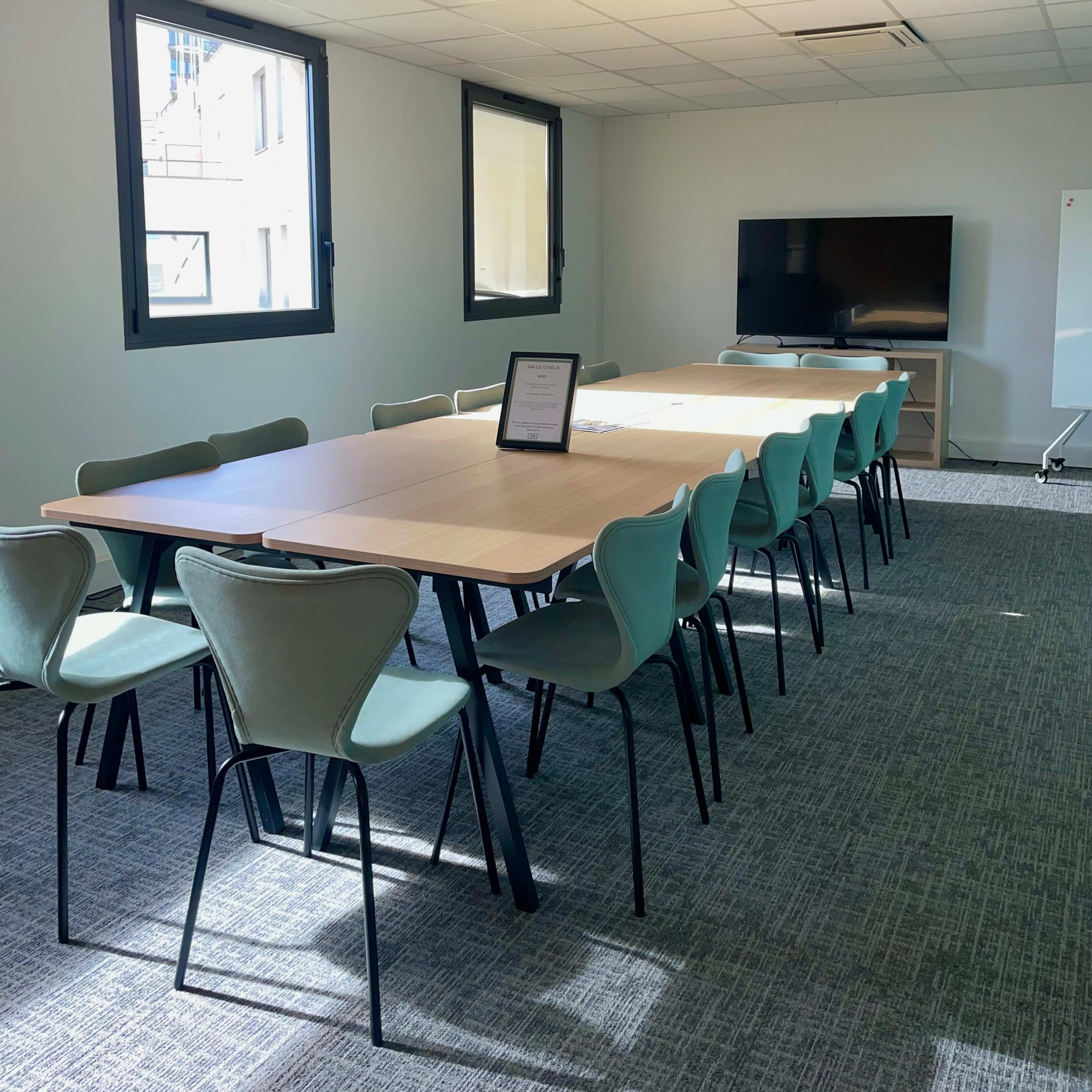A long conference table with teal chairs is positioned in a well-lit meeting room featuring a large TV and a whiteboard.