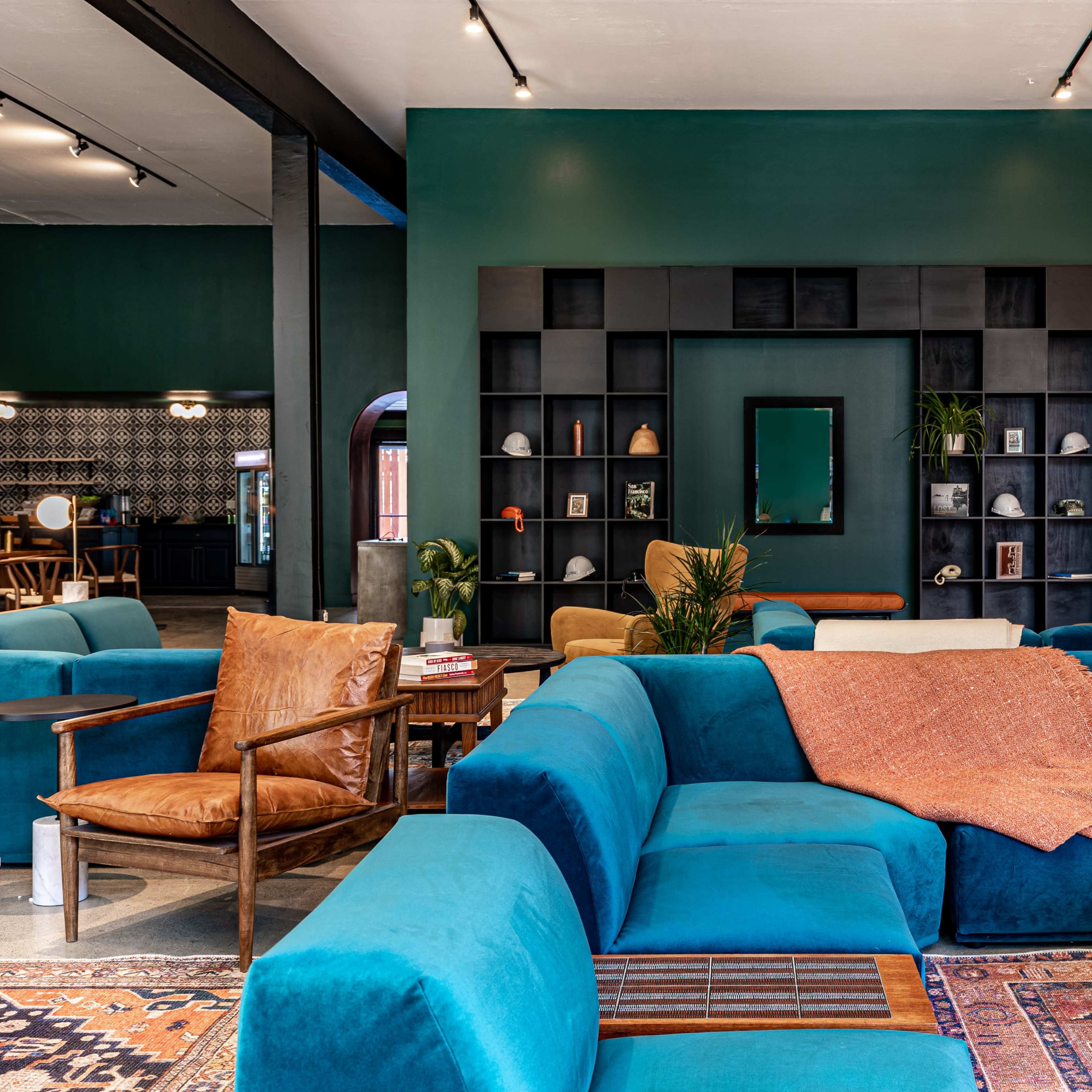 A spacious, stylish lounge area features a large teal sectional sofa, wooden accents, and decorative shelves against a deep green wall.