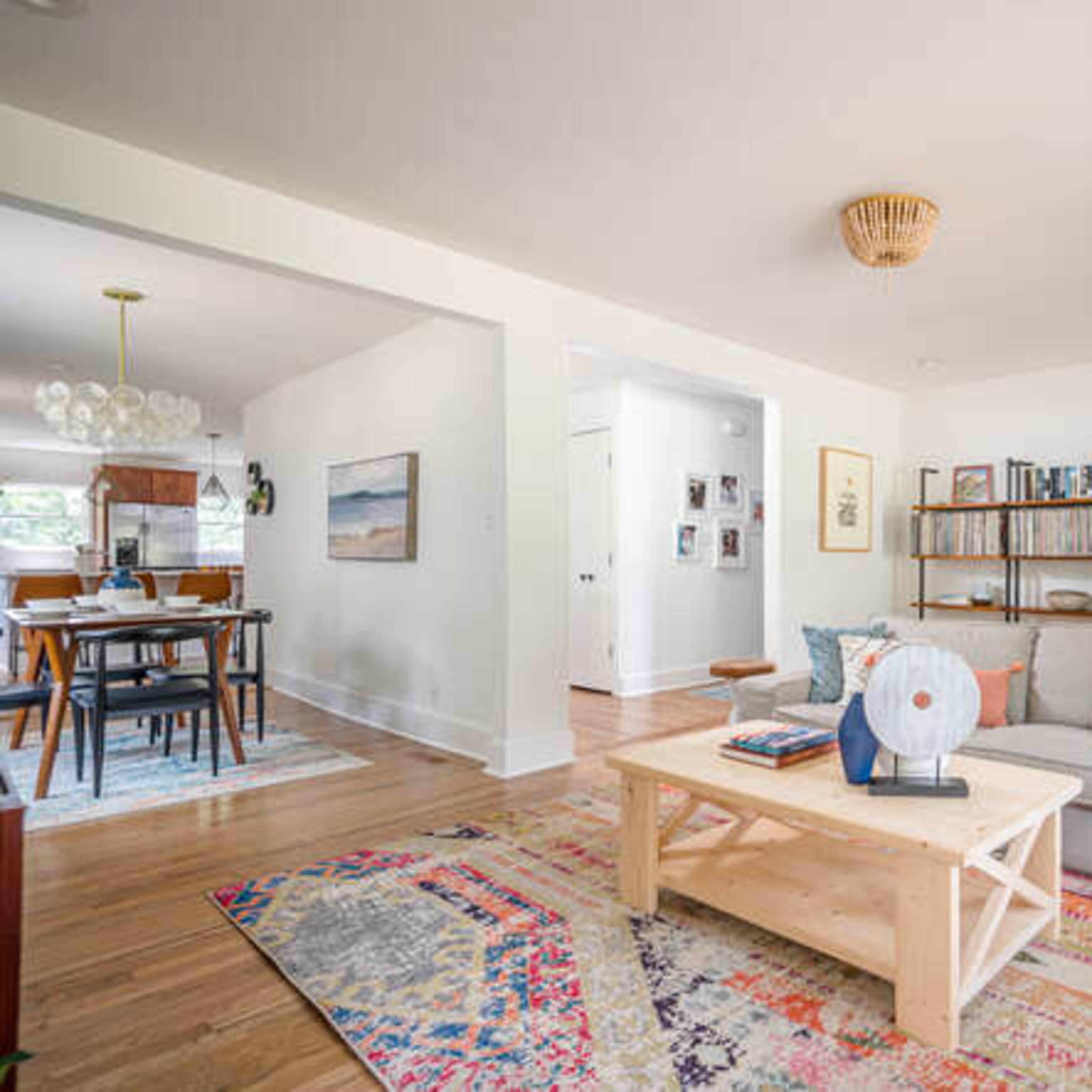 A cozy living room features a light gray sofa, a wooden coffee table, and a colorful area rug, with a bookshelf in the background and a dining area visible through an adjacent doorway.
