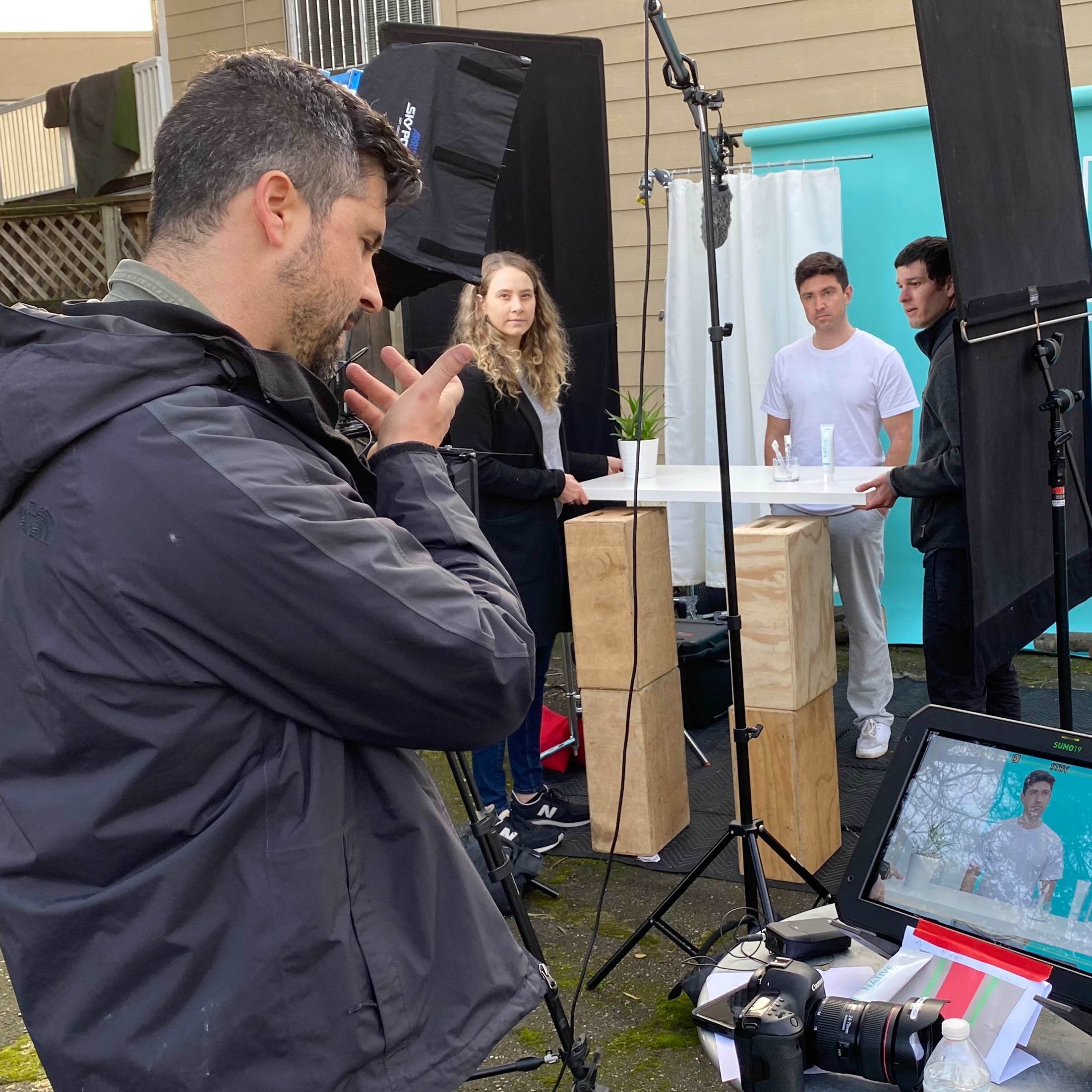 A photographer sets up a photoshoot with three models in front of a turquoise backdrop, surrounded by lighting equipment and a monitor displaying the shot.