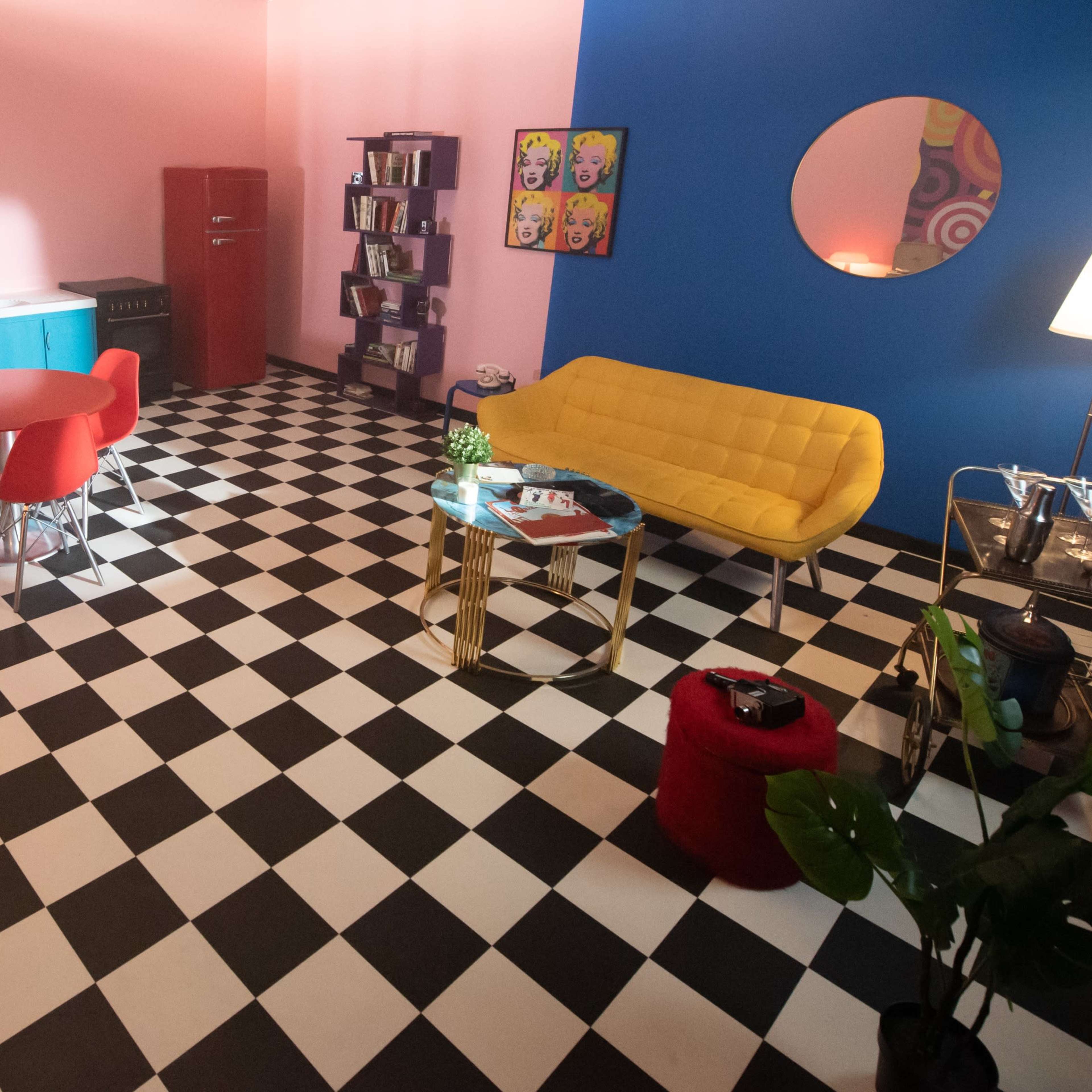 A brightly colored room with a checkered black-and-white floor featuring a blue accent wall, a yellow couch, red dining chairs, and a small kitchenette.