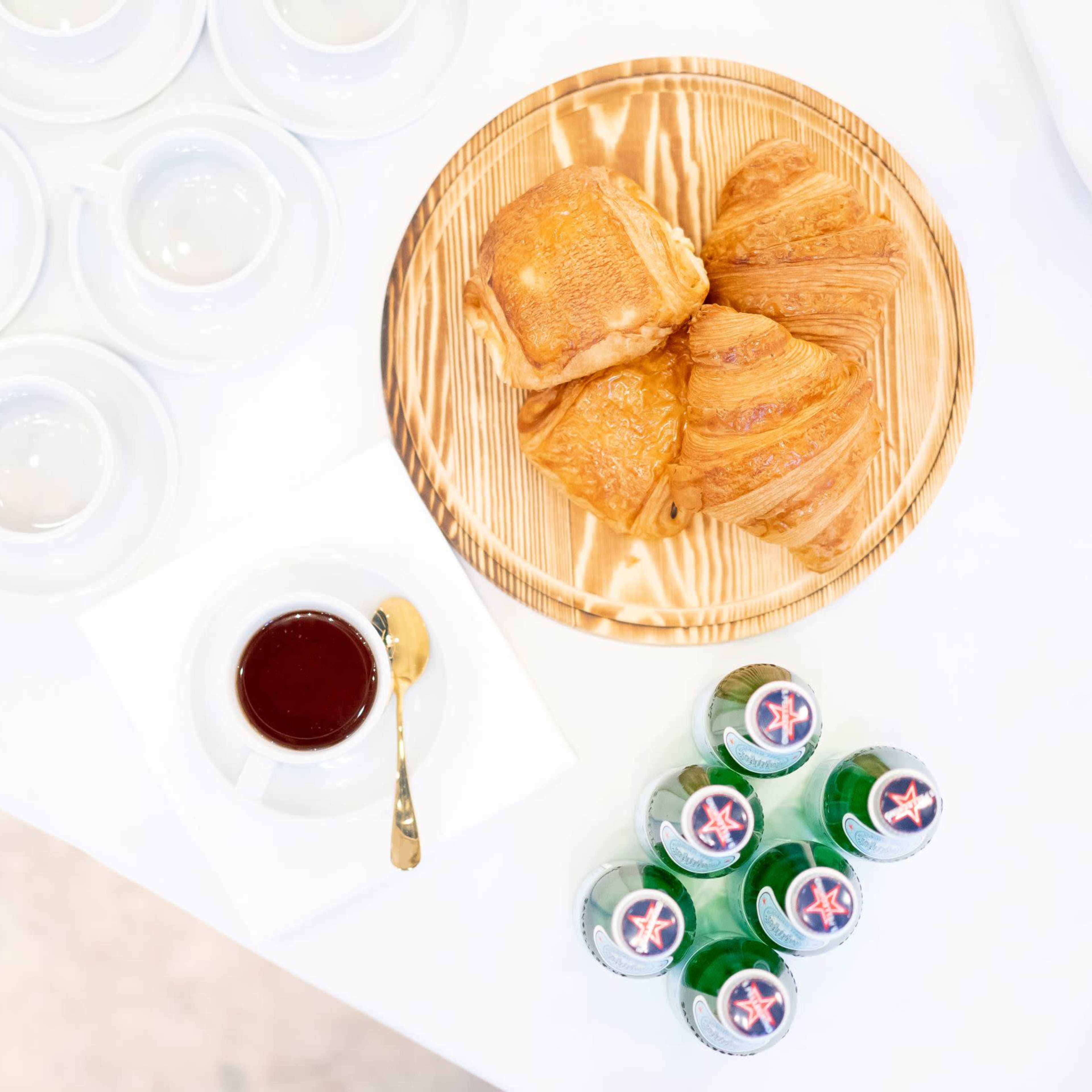 A wooden platter holds three croissants beside a cup of tea and several small plates, with a group of bottled beverages nearby.