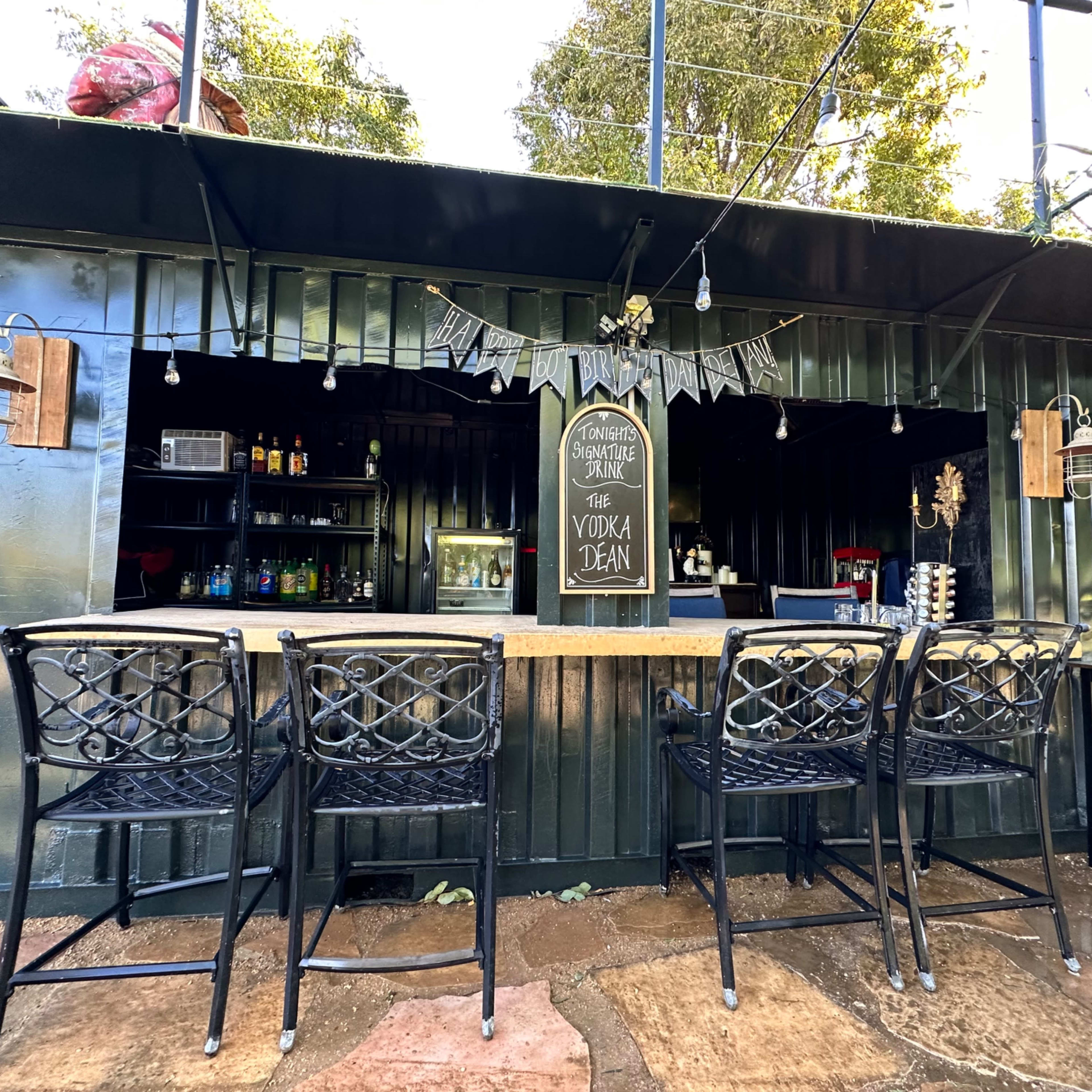 An outdoor bar area with a counter, several black metal stools, and string lights hanging above.