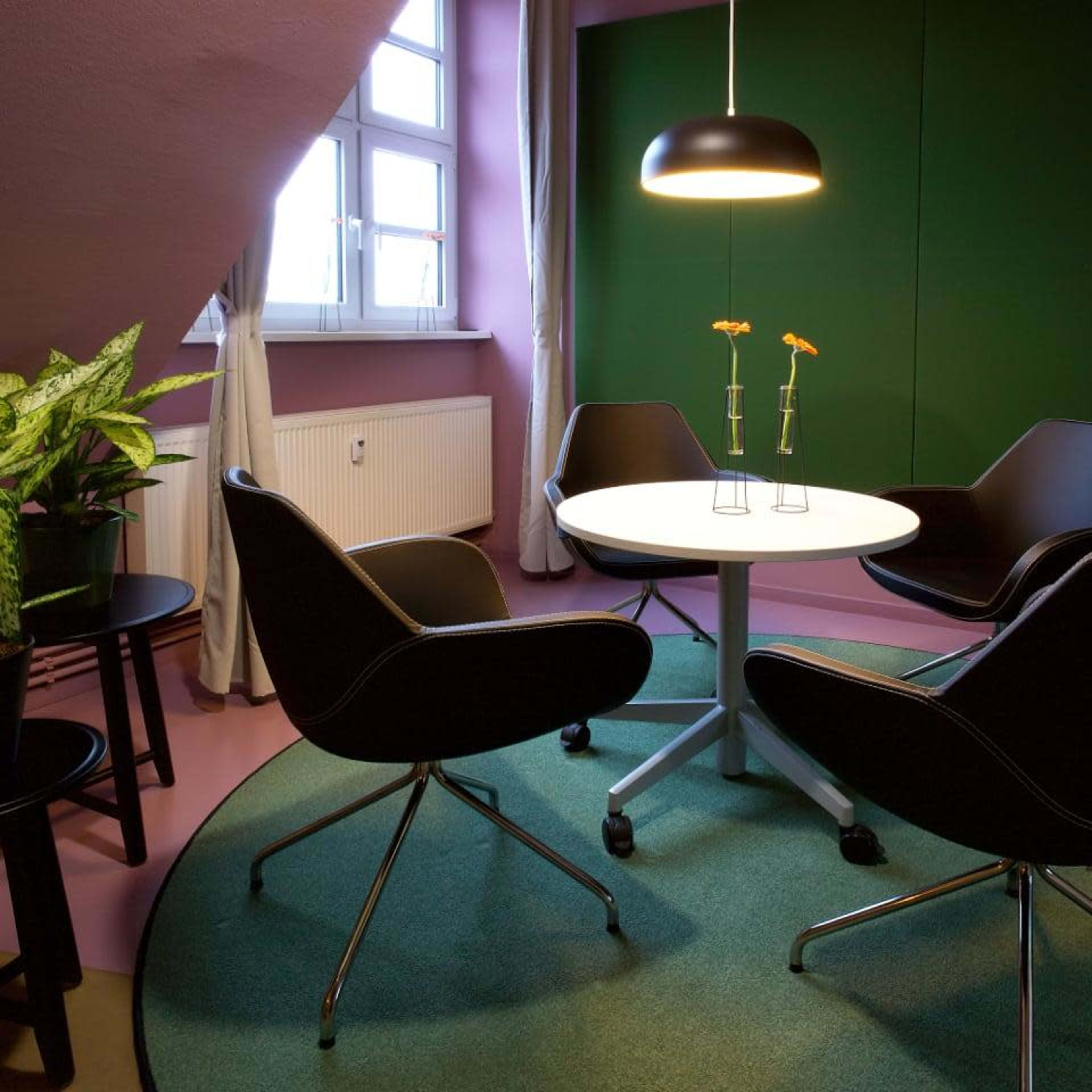 A small meeting room features a round table surrounded by four modern chairs, with a potted plant nearby and a large window letting in natural light.
