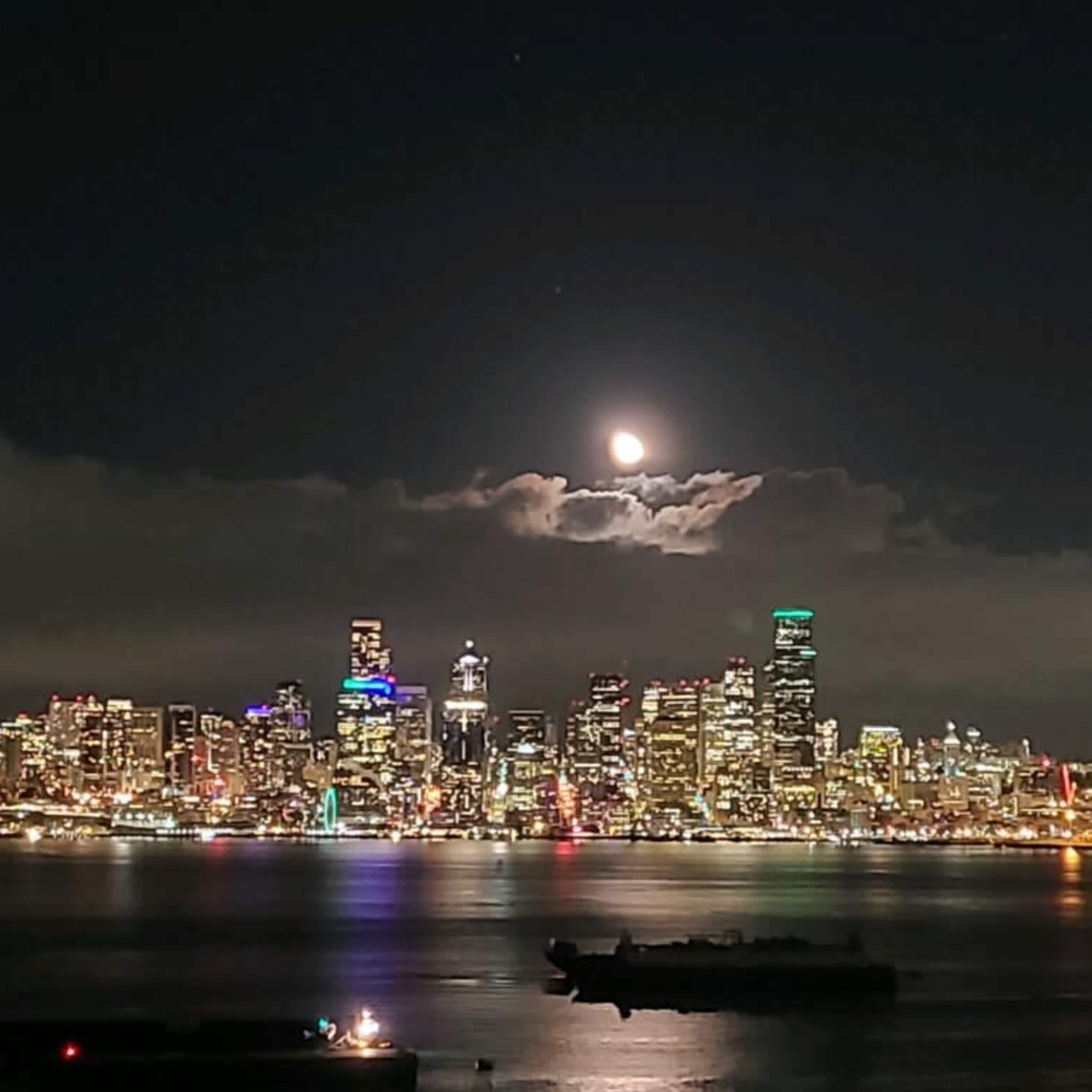 A full moon rises behind clouds over a shimmering cityscape at night, with a ship in the foreground.