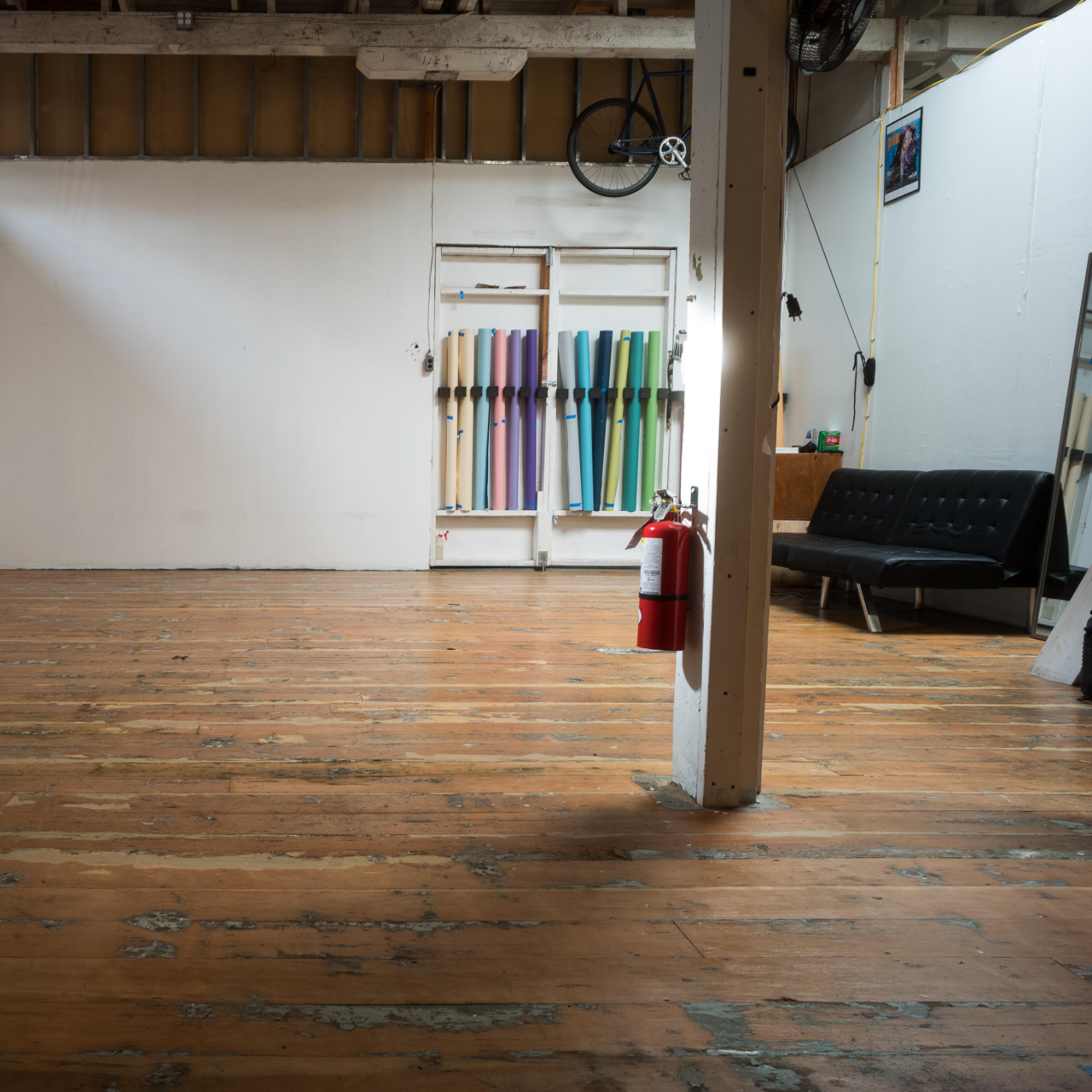 An empty room features wooden floors, a white painted wall, a couch, a fire extinguisher, and colorful materials hanging on another wall.