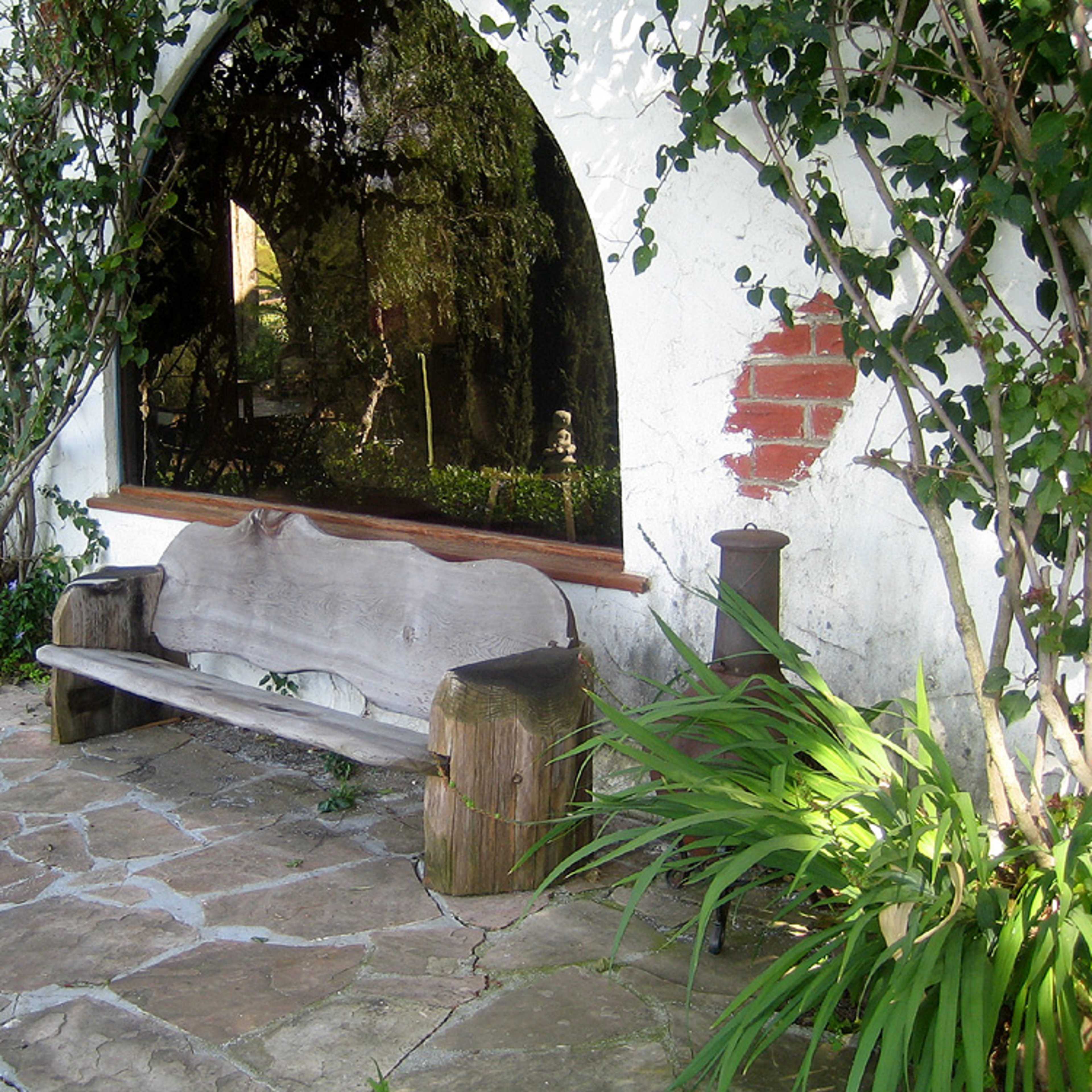 A wooden bench sits against a wall surrounded by greenery and stone pathways.