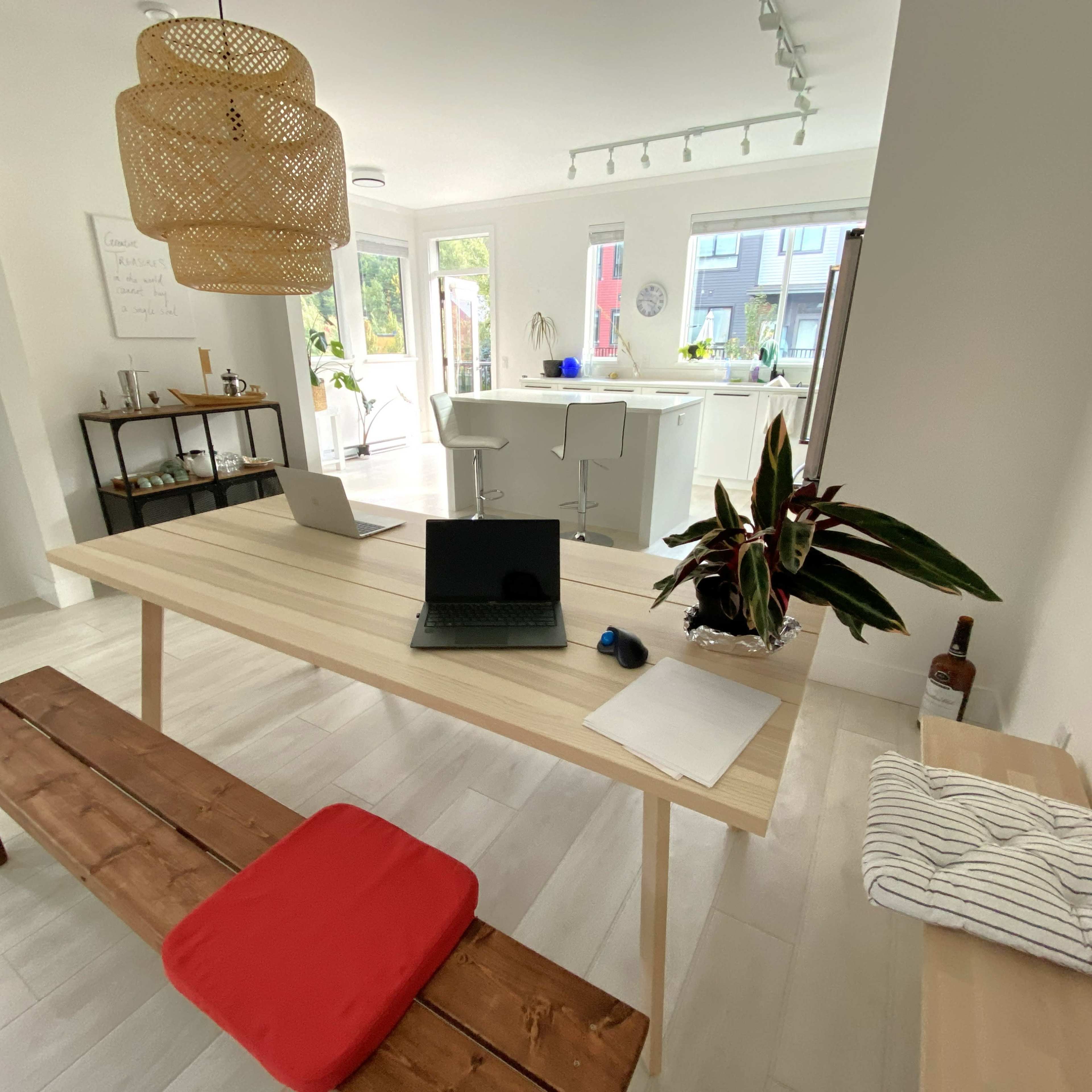 A modern interior space featuring a wooden table with a laptop, a seating bench with a red cushion, and a bright kitchen area in the background.