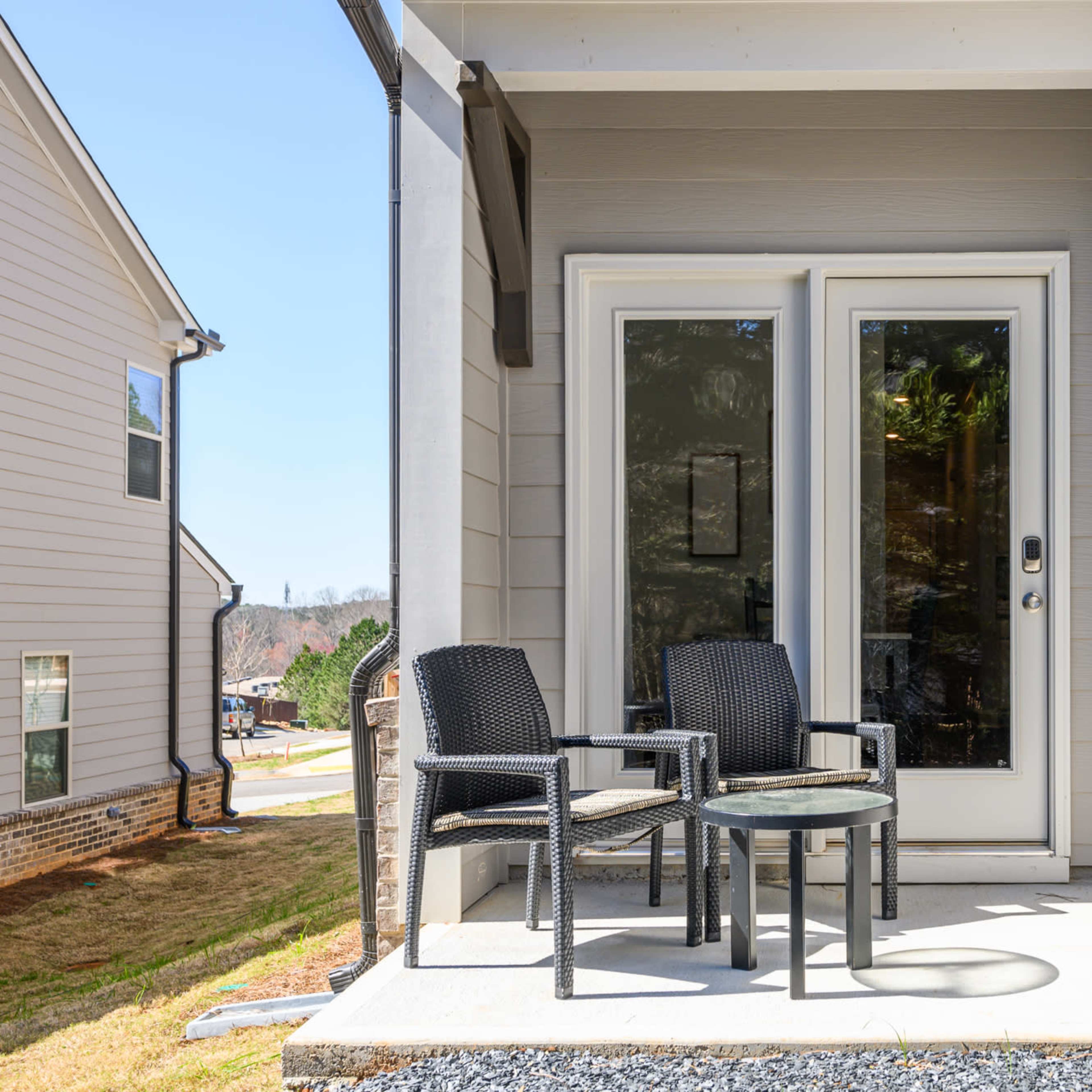 A small patio area with two black chairs and a glass table in front of double glass doors.