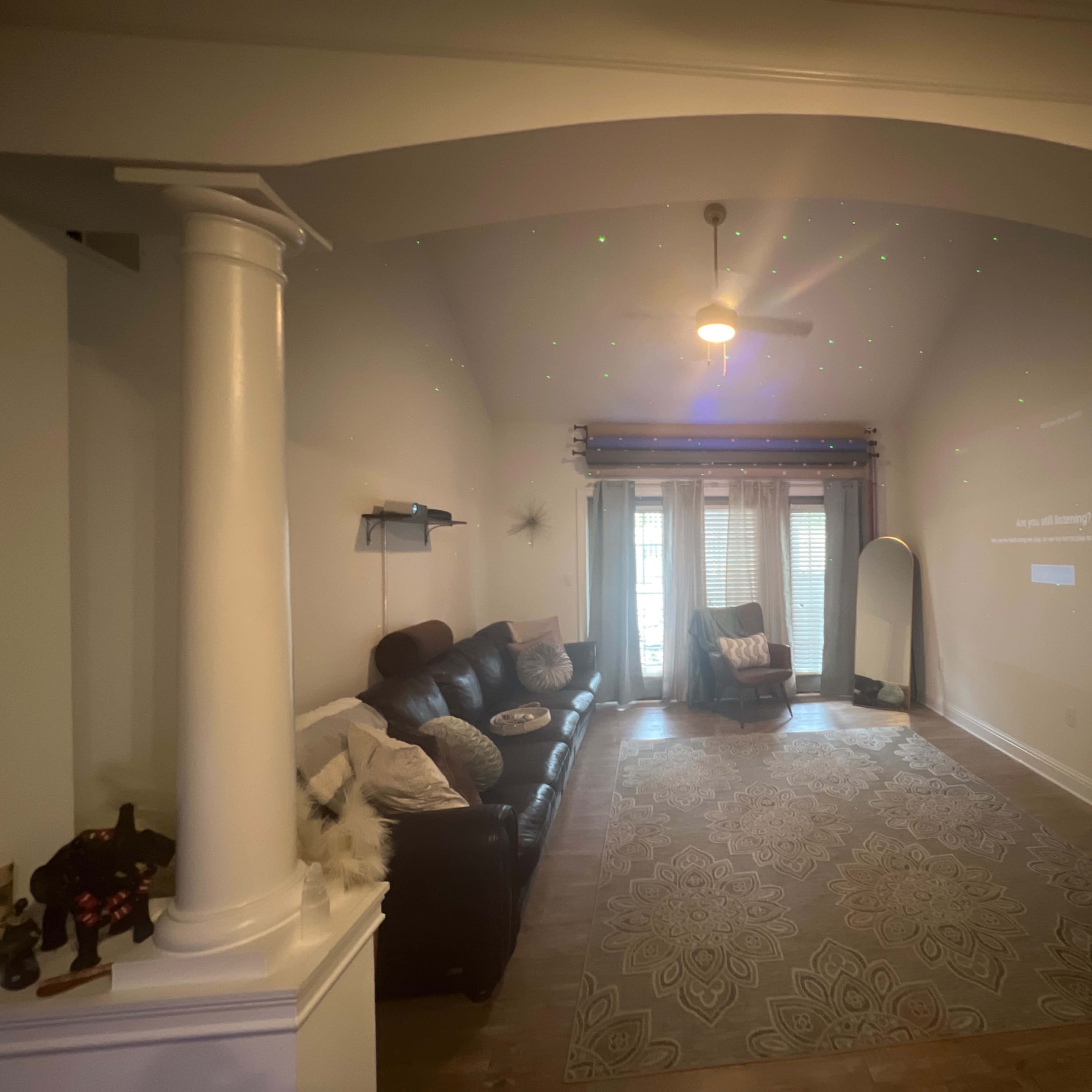 The living room features a leather sofa, a mirror, and decorative lighting projected on the wall, with large windows draped in sheer curtains.