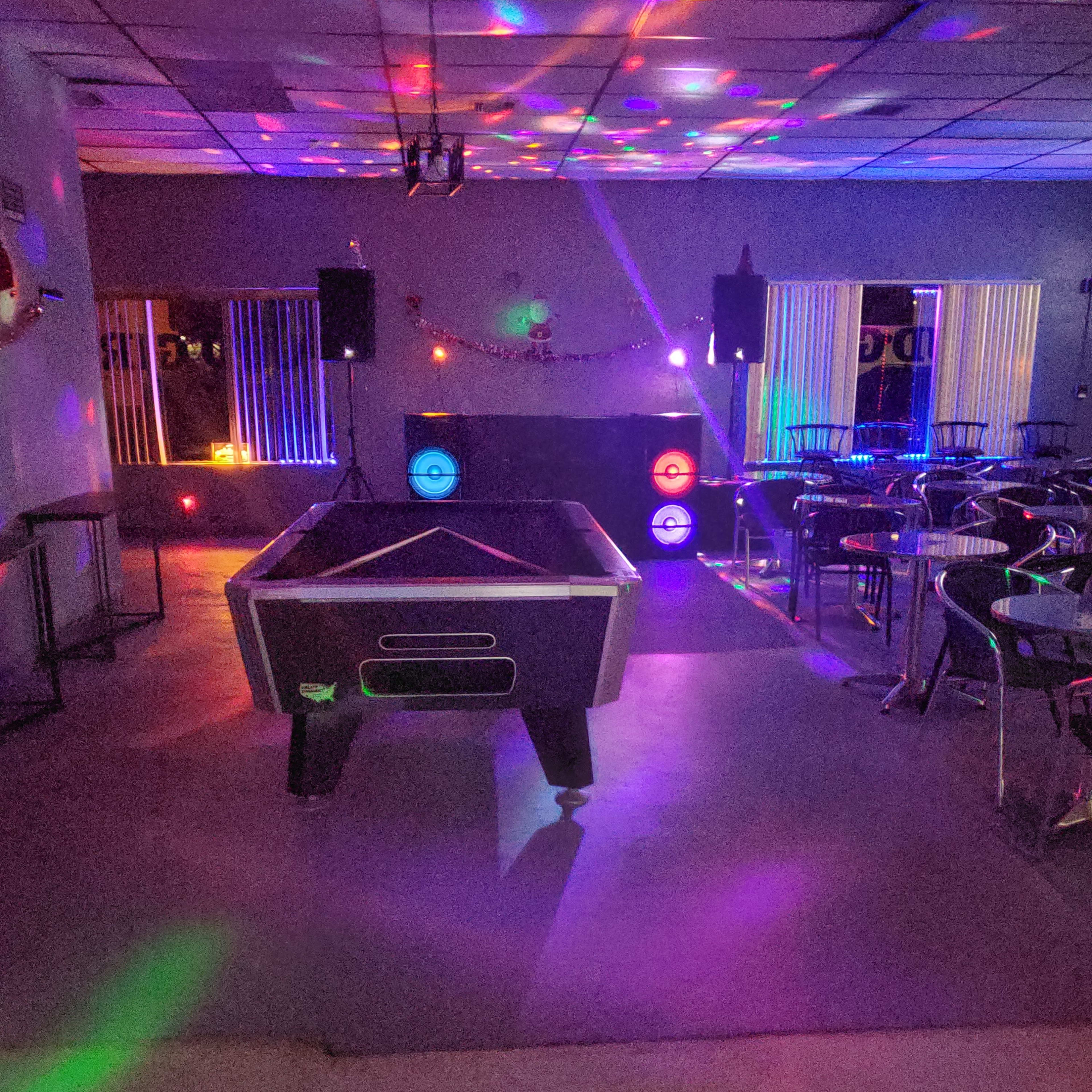 A dimly lit recreational room with a pool table at the center, surrounded by colorful lights and a small stage area with speakers.