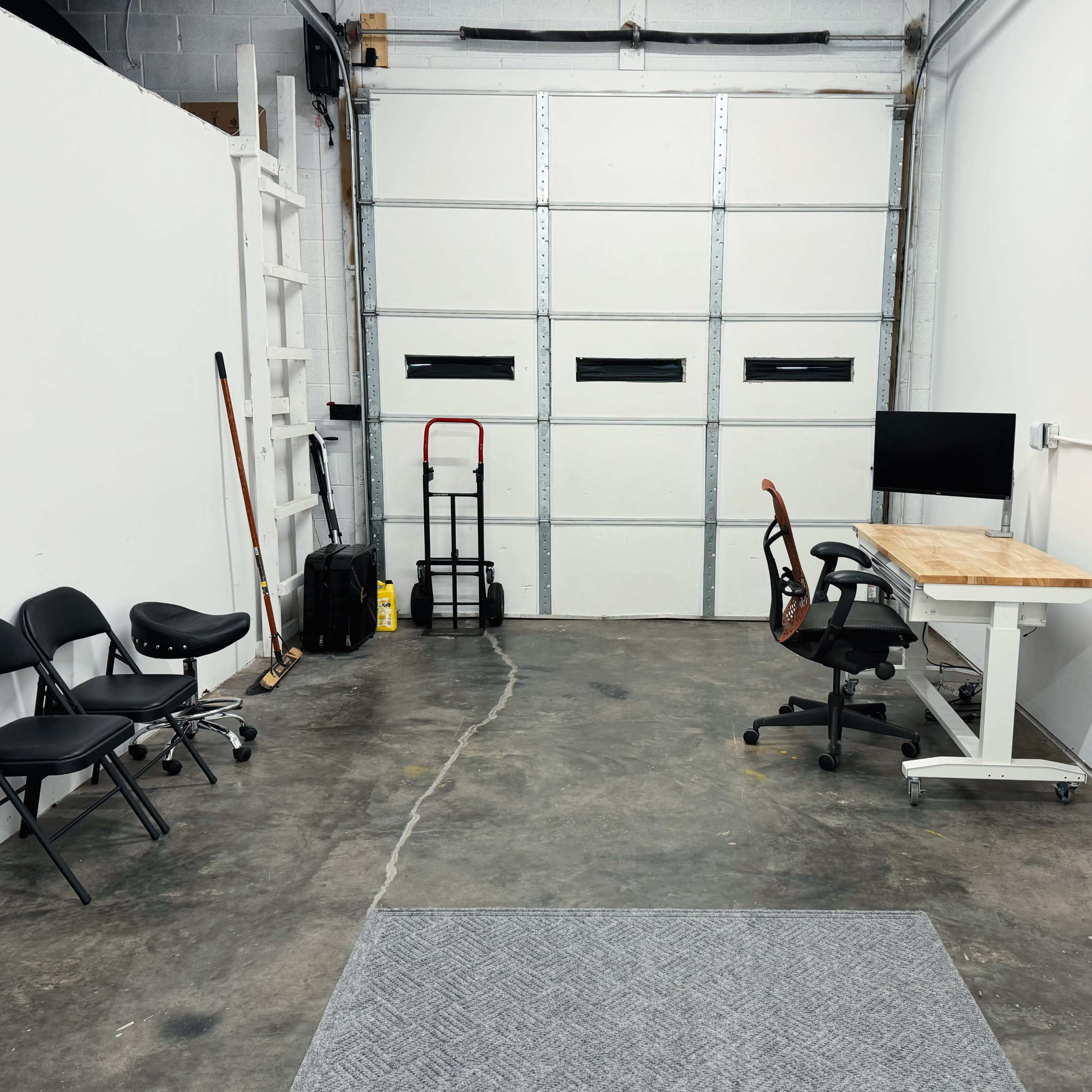 A sparse workspace with two chairs and a stool positioned next to a desk with a computer, opposite a garage door and additional empty space.