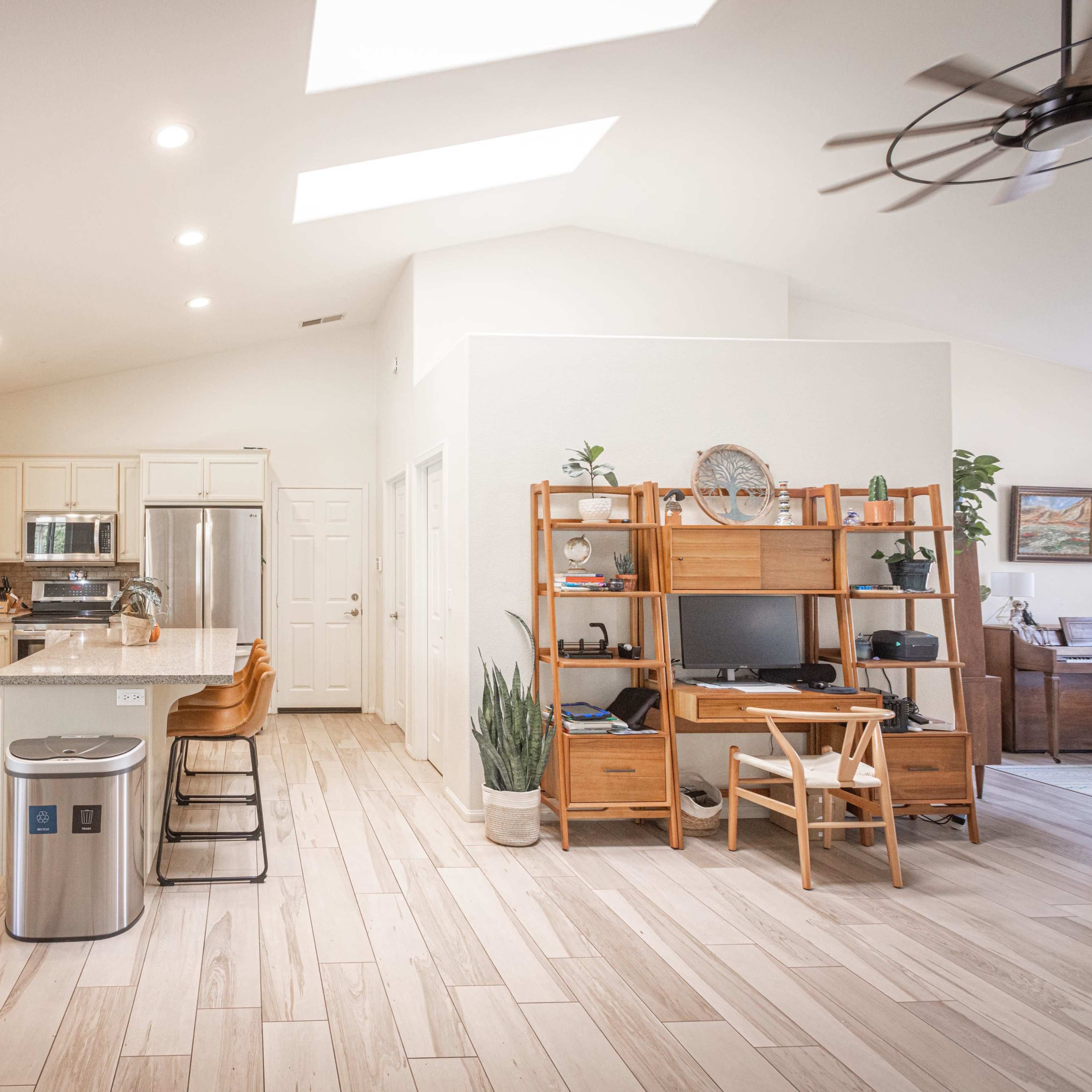 A modern kitchen and living area with light wood flooring, equipped with stainless steel appliances, a dining table, and a built-in desk.