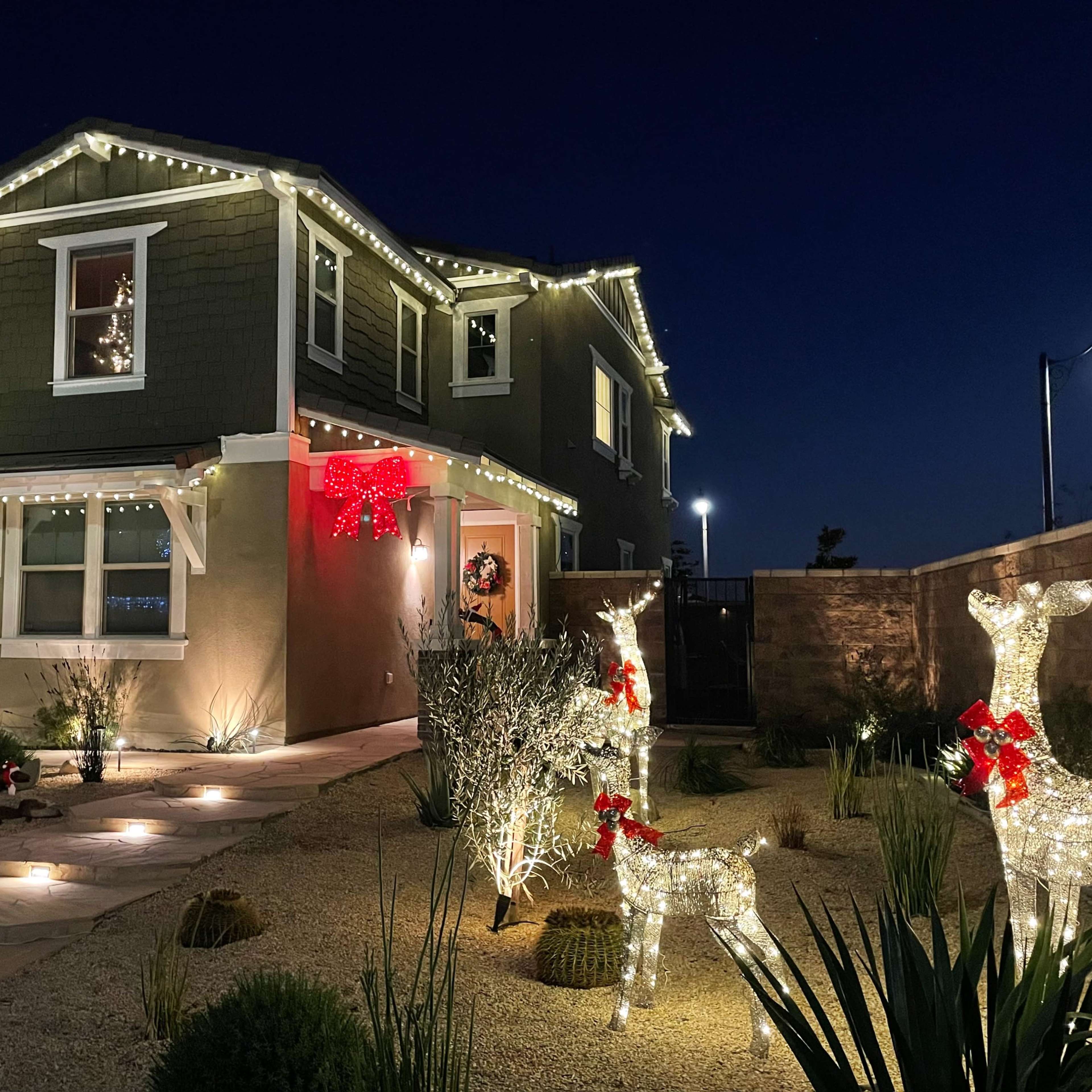 A two-story house is adorned with Christmas lights and decorations, featuring illuminated reindeer in the front yard and a warmly lit pathway.