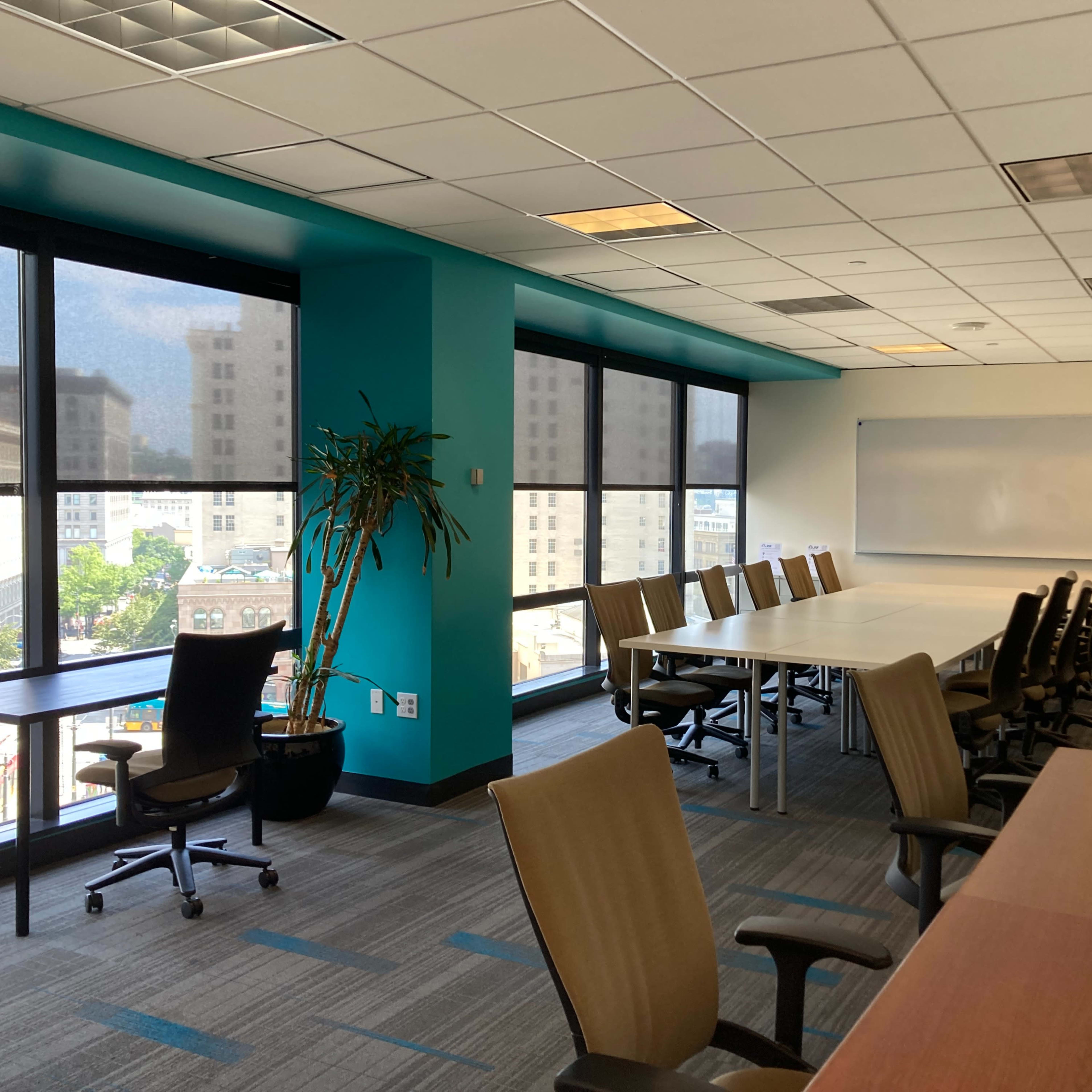 A conference room with large windows, a long table, and multiple chairs is shown, alongside a desk and greenery.