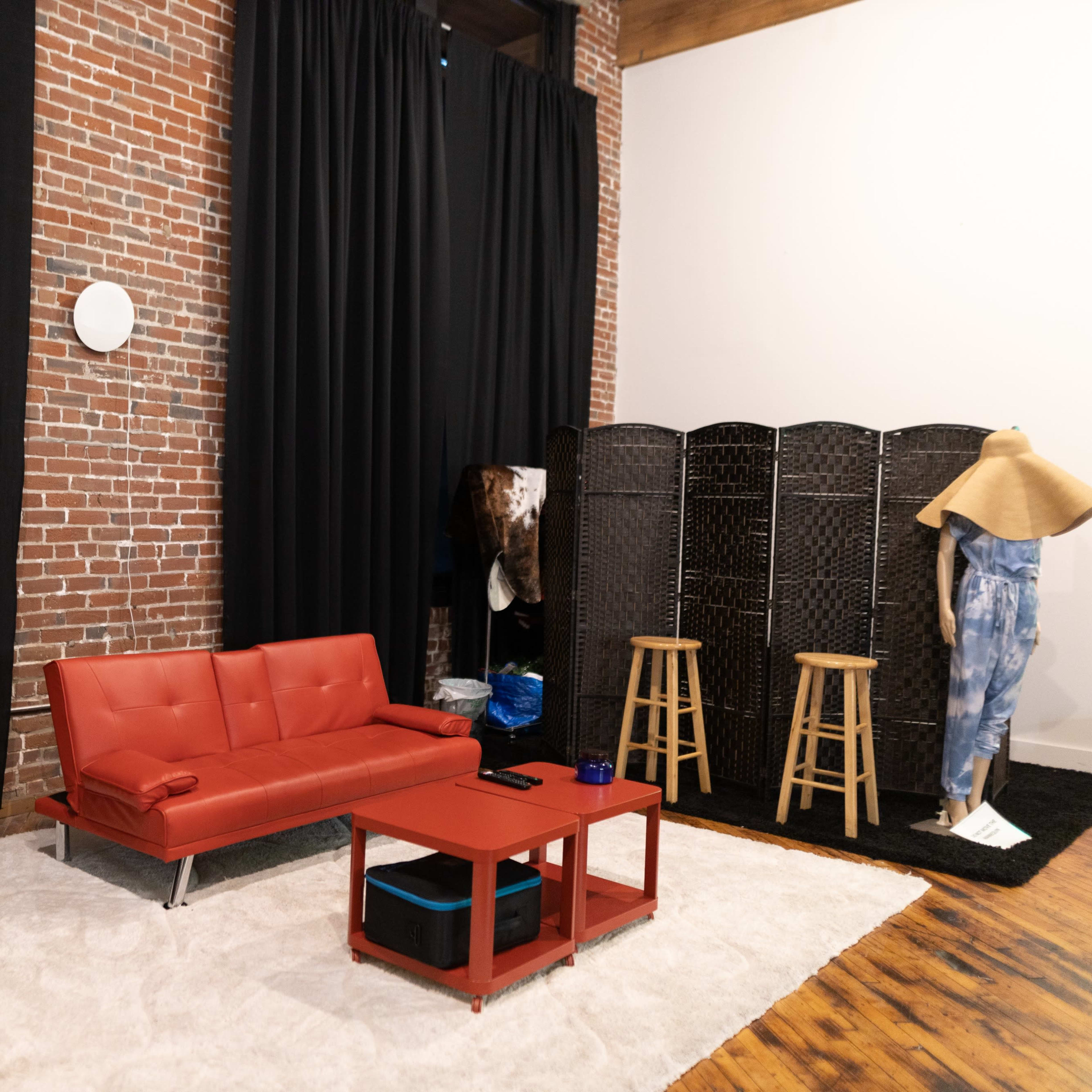 A contemporary living space with a red sofa, a hanging bubble chair, a coffee table, a folding screen, and two wooden stools against a backdrop of exposed brick and black curtains.