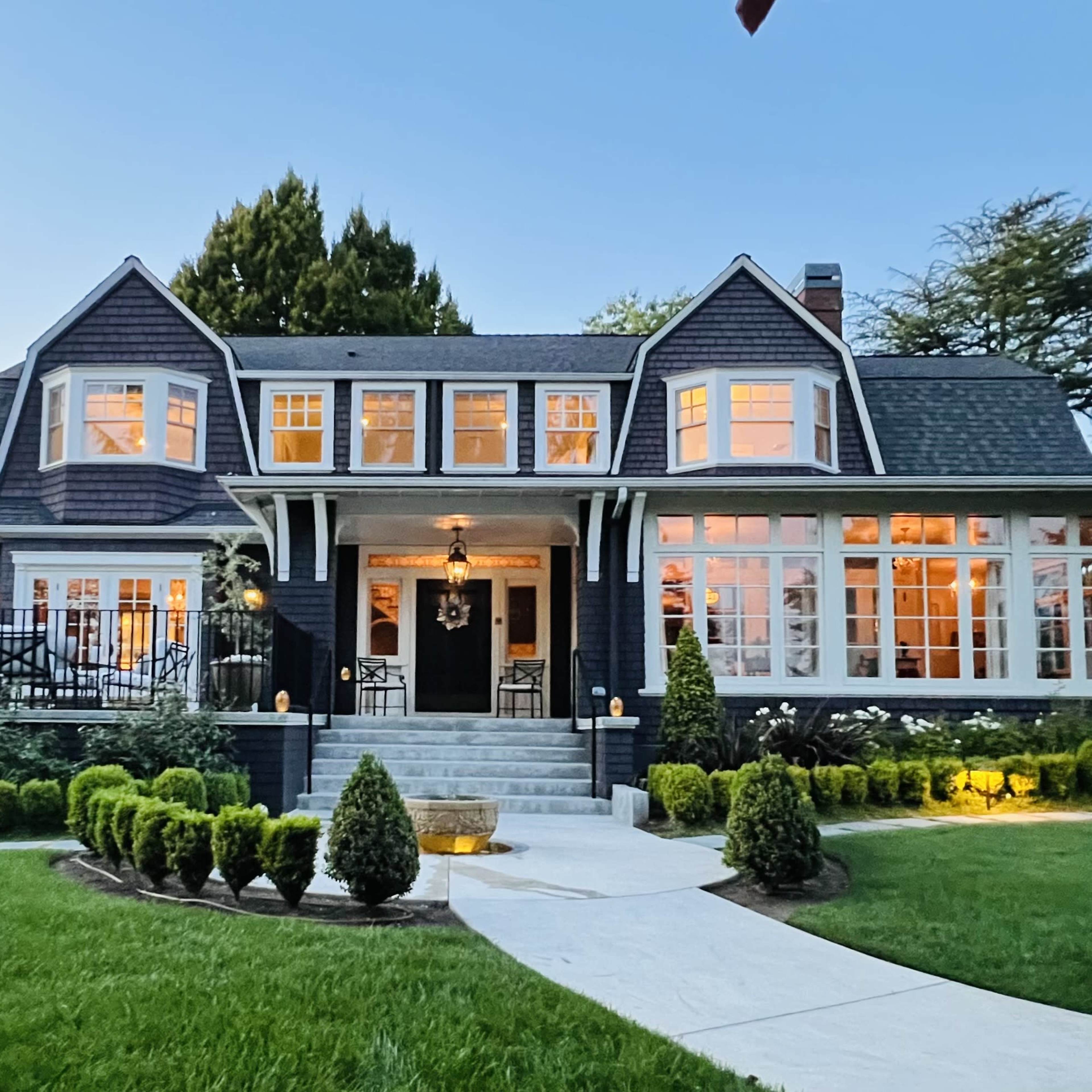 A large, two-story house with multiple gabled roofs, numerous windows, and a well-manicured lawn featuring symmetrical hedges and pathway lighting.
