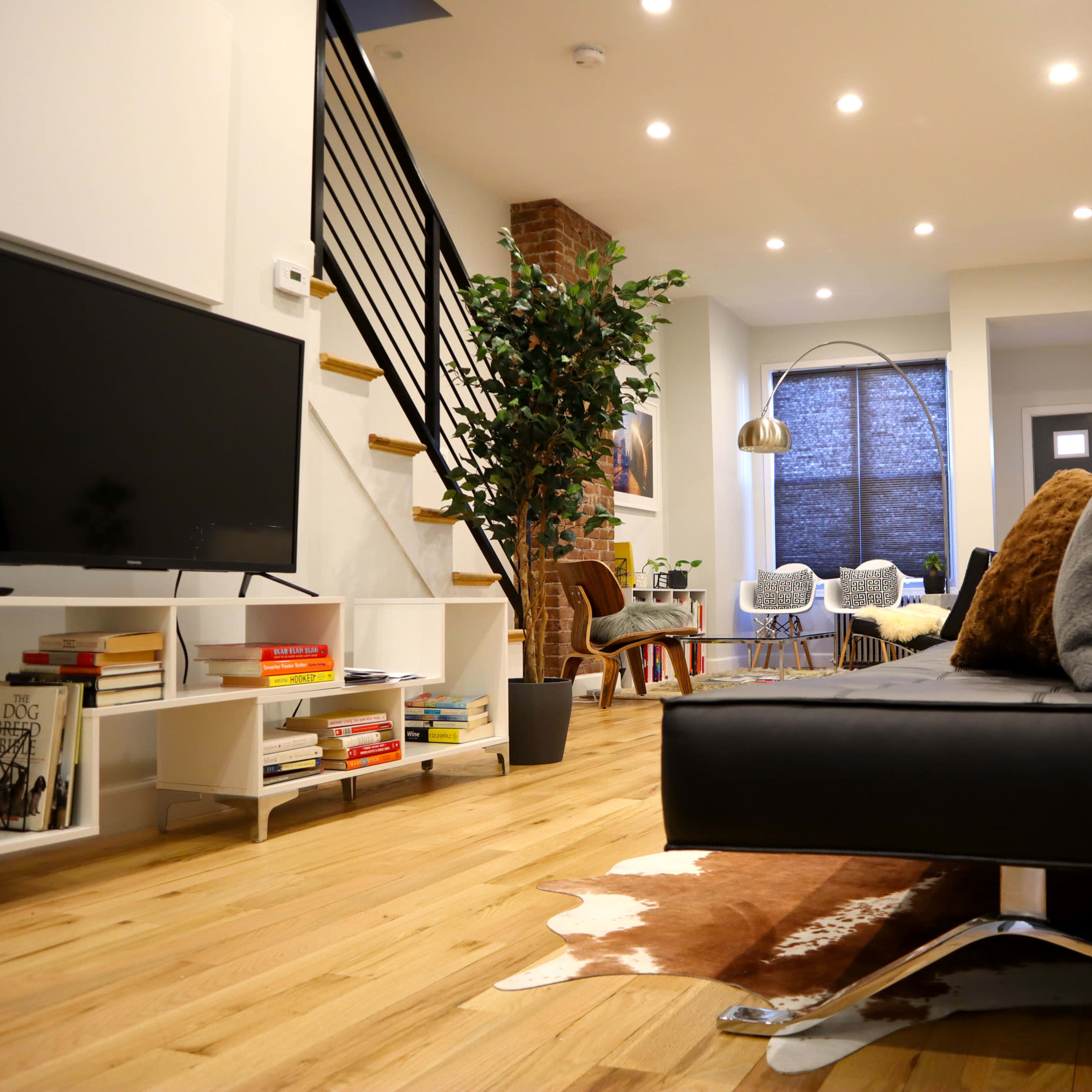 The scene features a modern living room with a black sofa, a television on a white shelf, a potted plant, and a staircase that leads to an upper level.