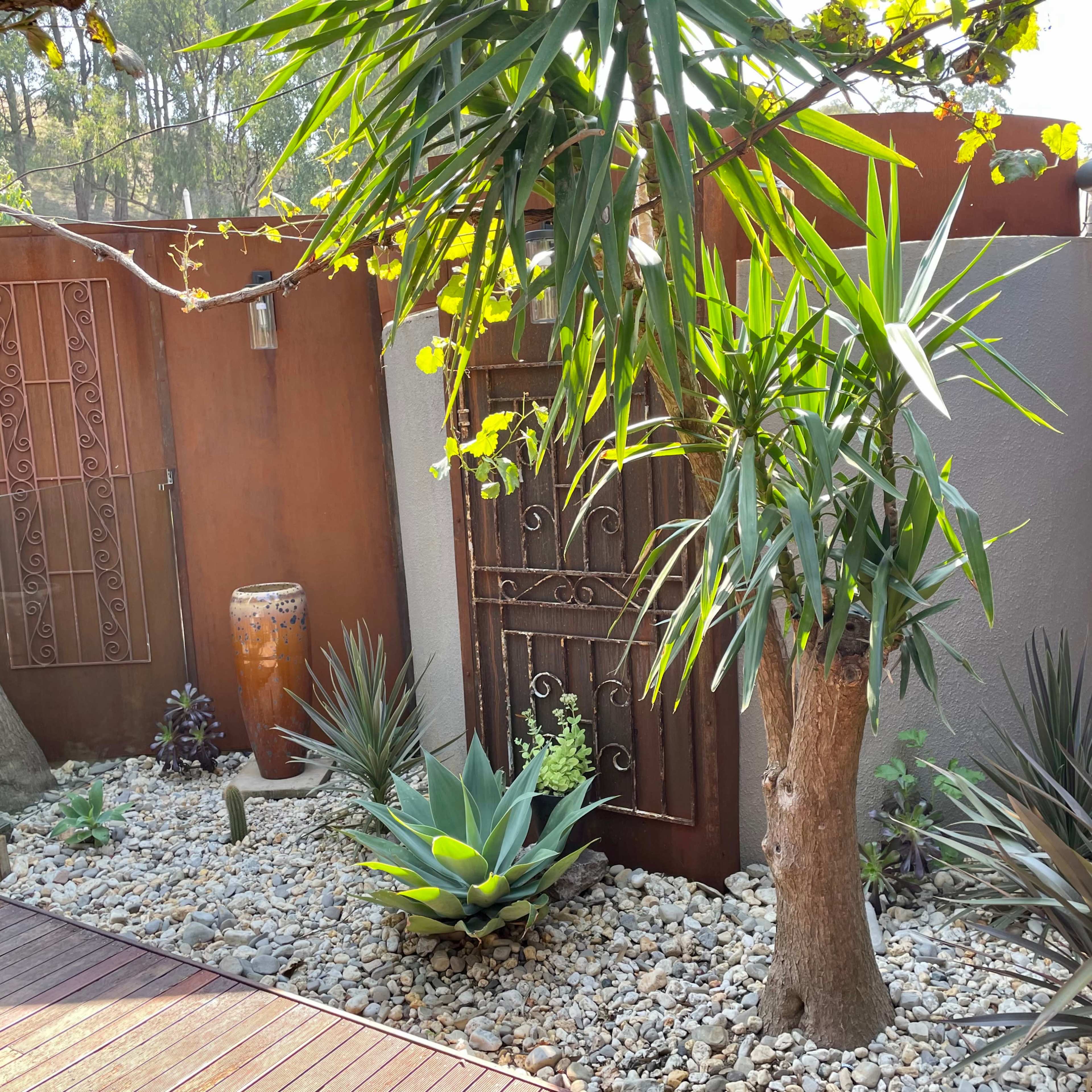 The scene features a landscaped area with various plants, a decorative wall, and a gated entrance surrounded by rocks.