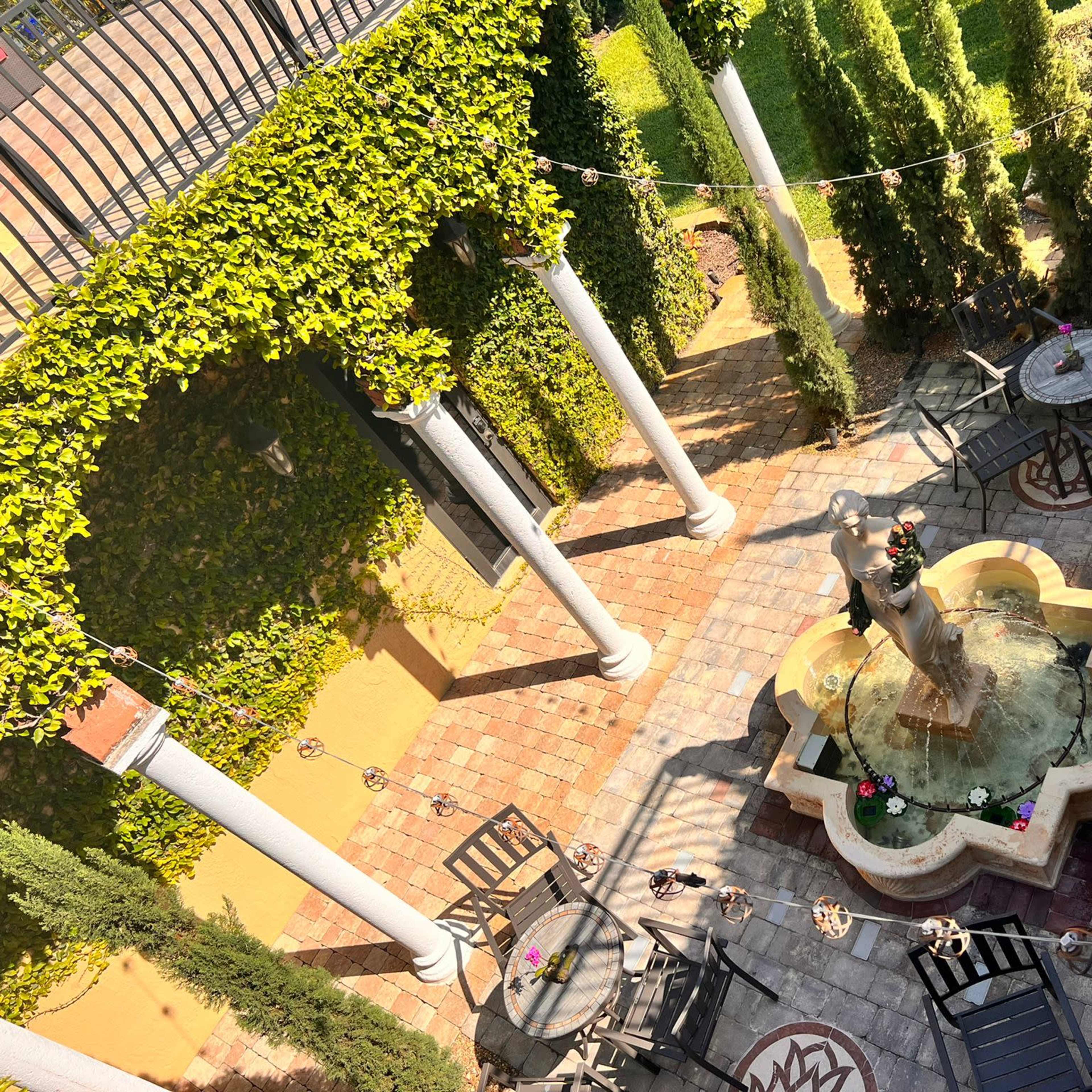 A courtyard with a central fountain surrounded by stone paving, tables, and chairs, encircled by greenery and climbing vines.