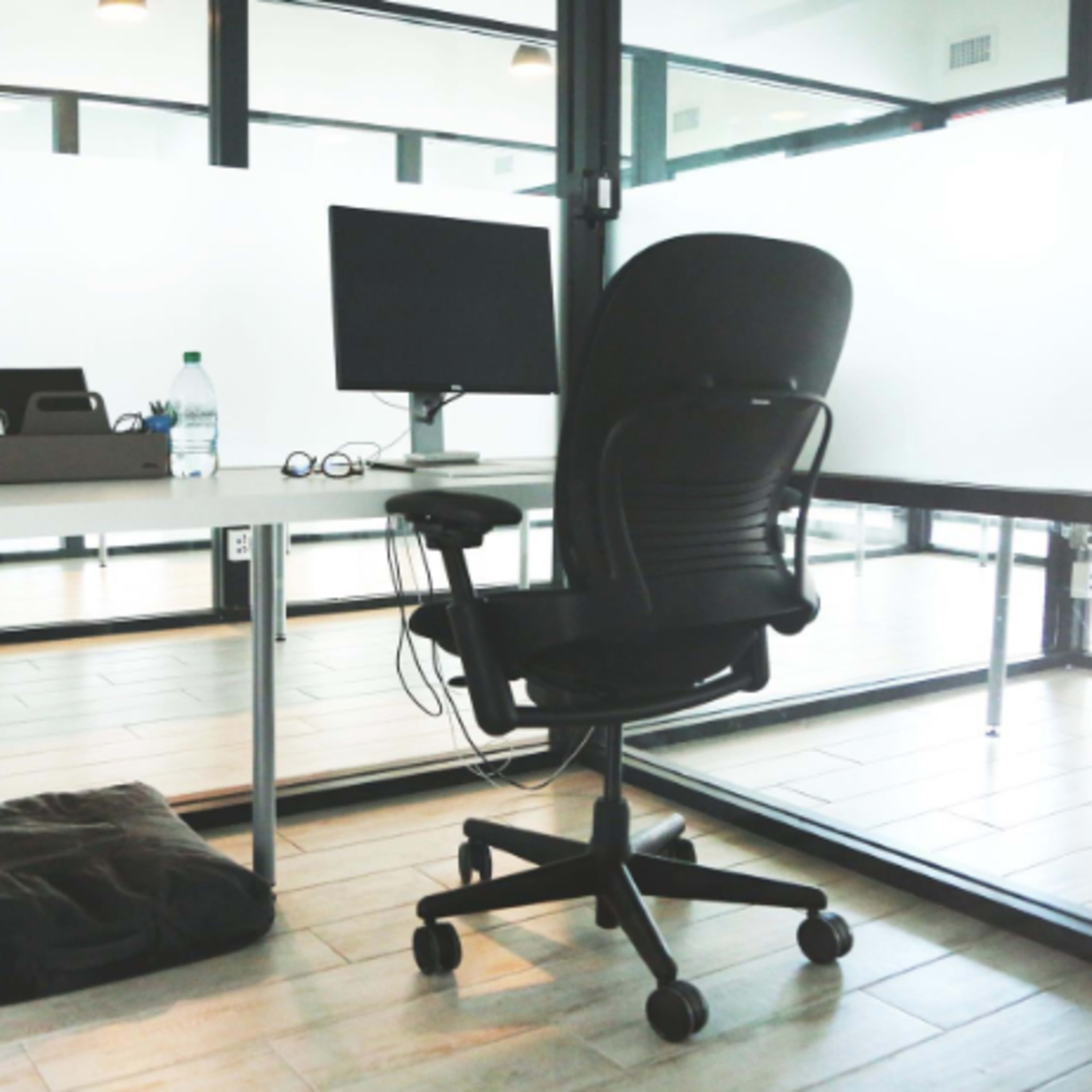 An office workspace featuring a desk with a computer monitor, a chair, a water bottle, glasses, and a potted plant, with a cushion and bag positioned on the floor nearby.