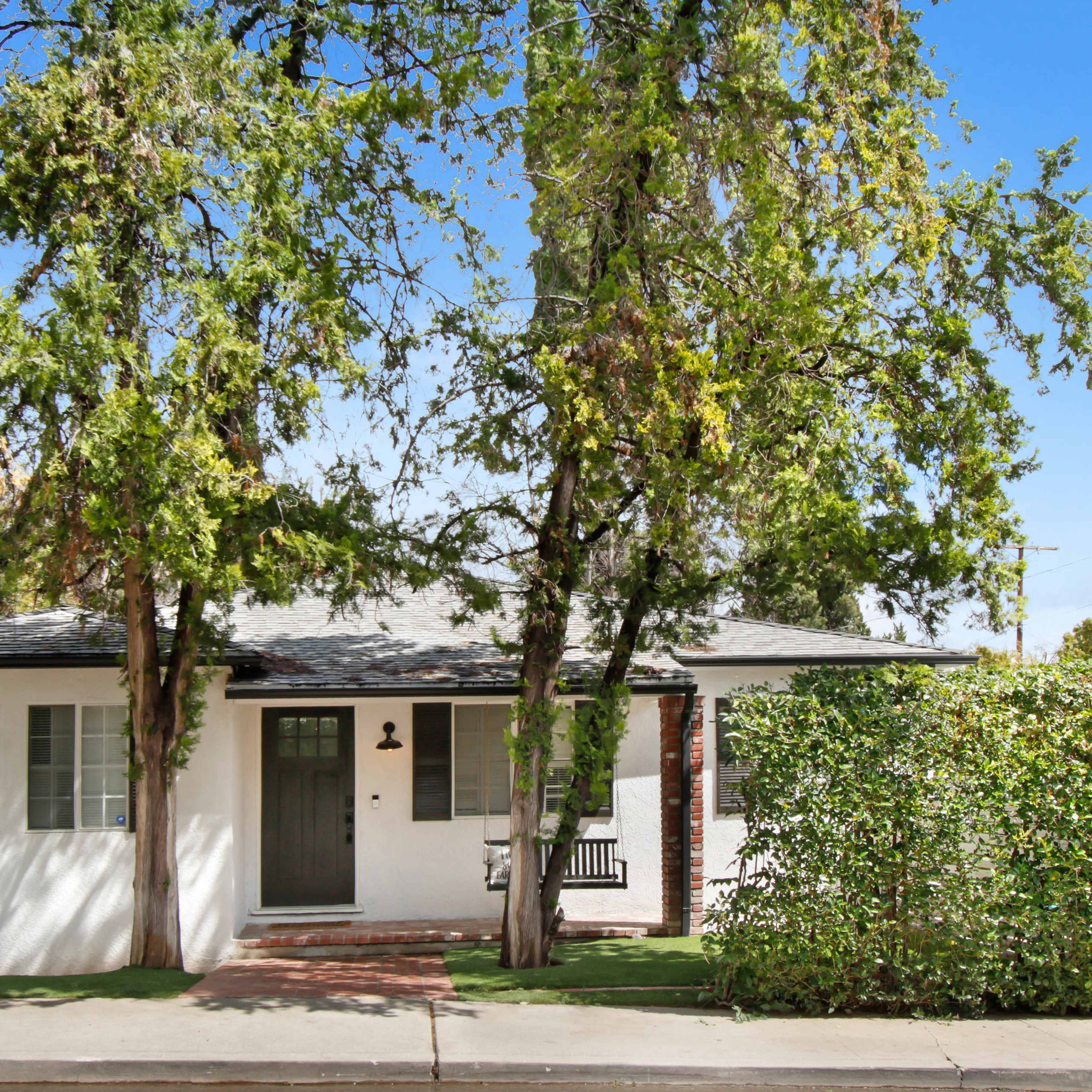 A single-story, white-painted house with green shutters is surrounded by trees and shrubs along a paved walkway.