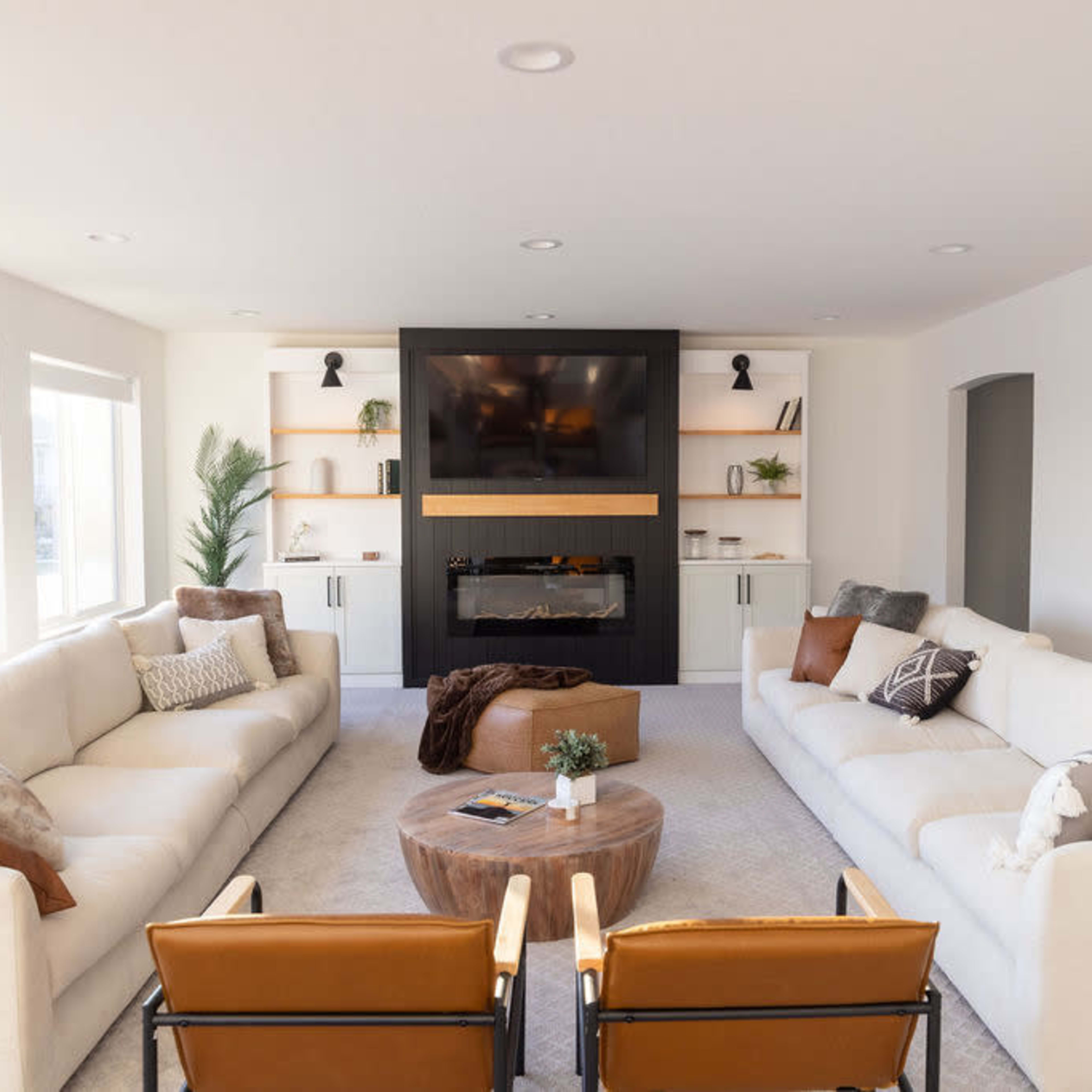 A modern living room with two symmetrical sofas, two wooden armchairs, a round coffee table, and a wall-mounted television above an electric fireplace.