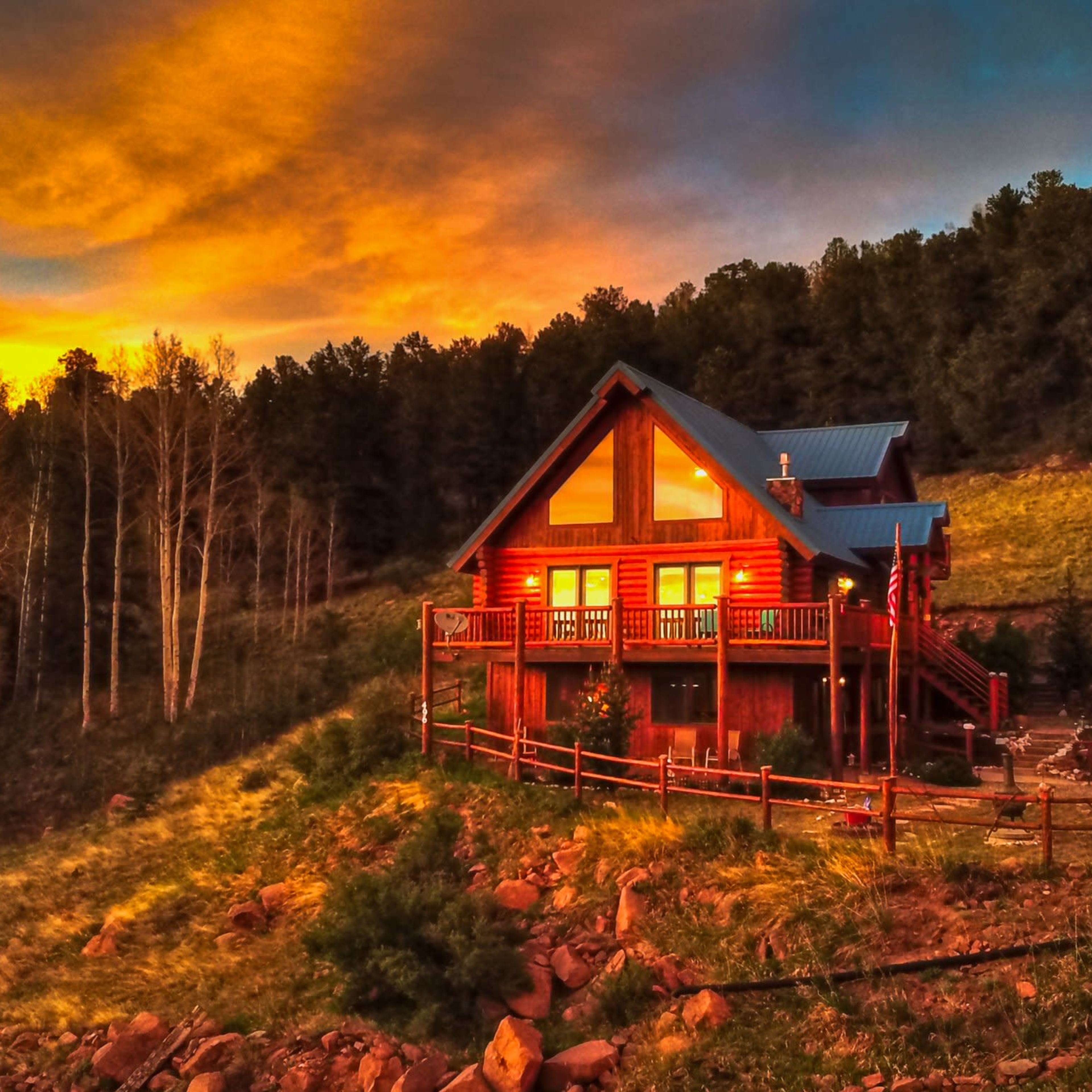 A wooden cabin with large windows is situated on a hillside surrounded by trees, under a colorful sunset sky.