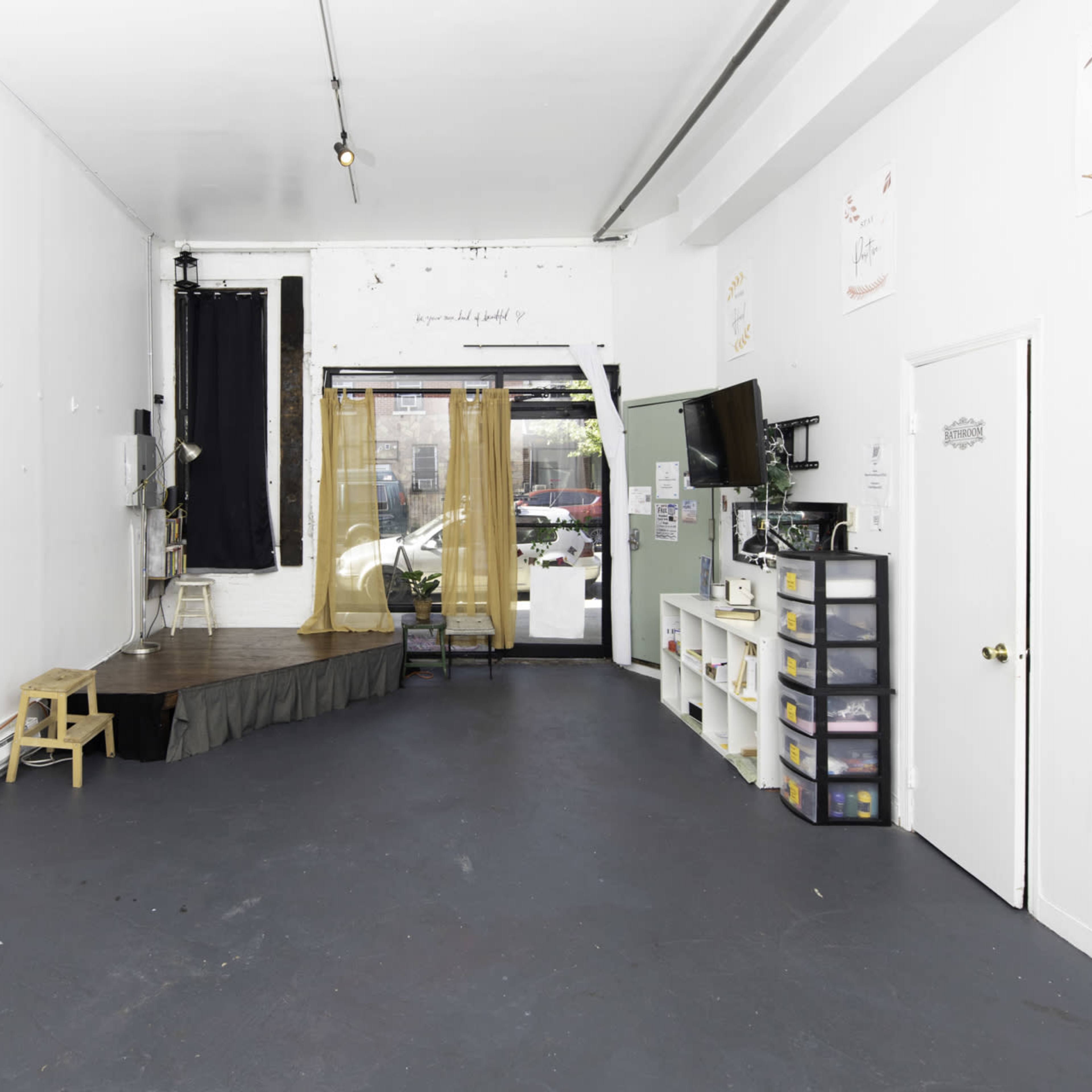 An empty room with a black floor, a small stage area, a television on the wall, and storage shelves along one side.