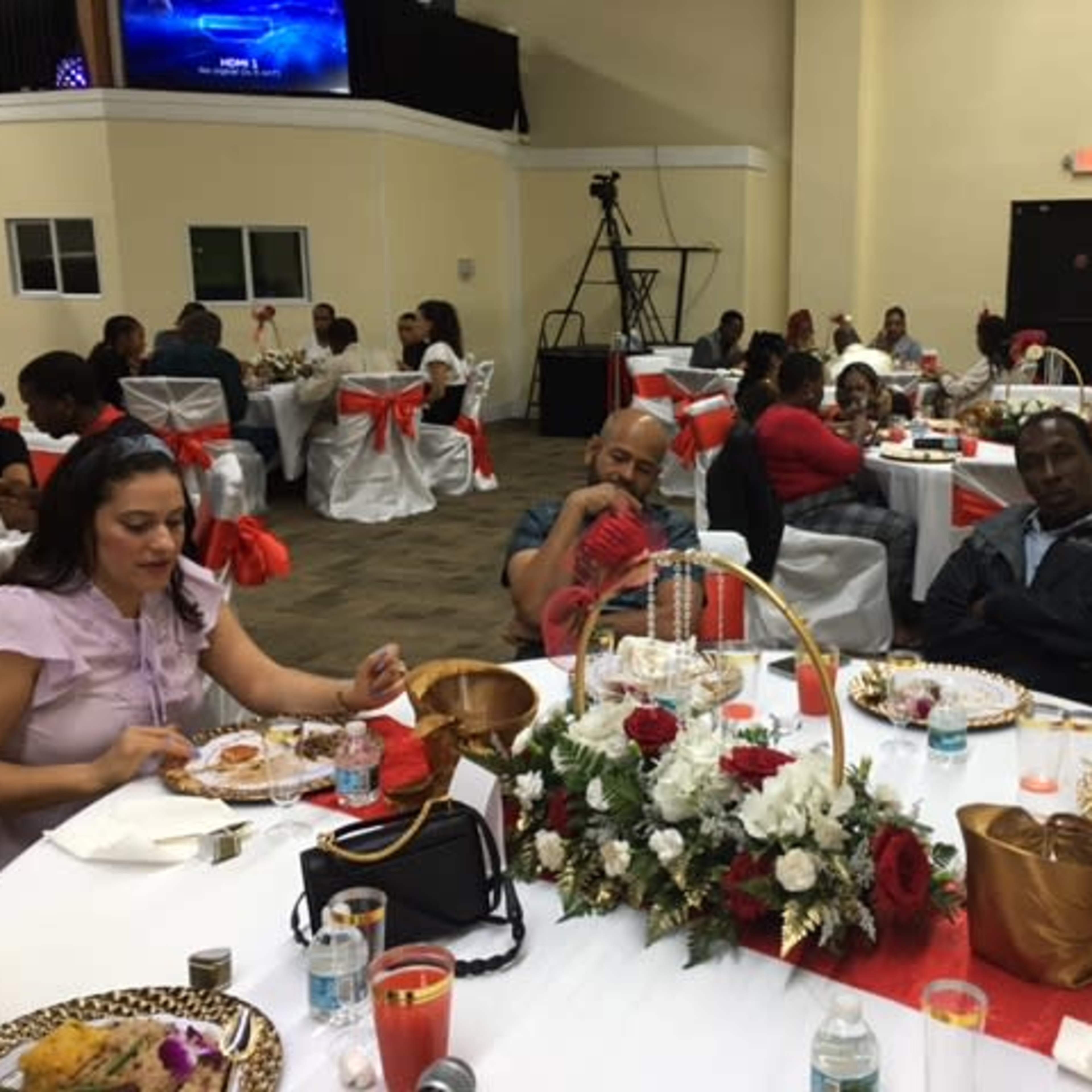 A banquet hall filled with guests seated at tables adorned with white tablecloths and red chair sashes, enjoying a meal in a festive atmosphere.