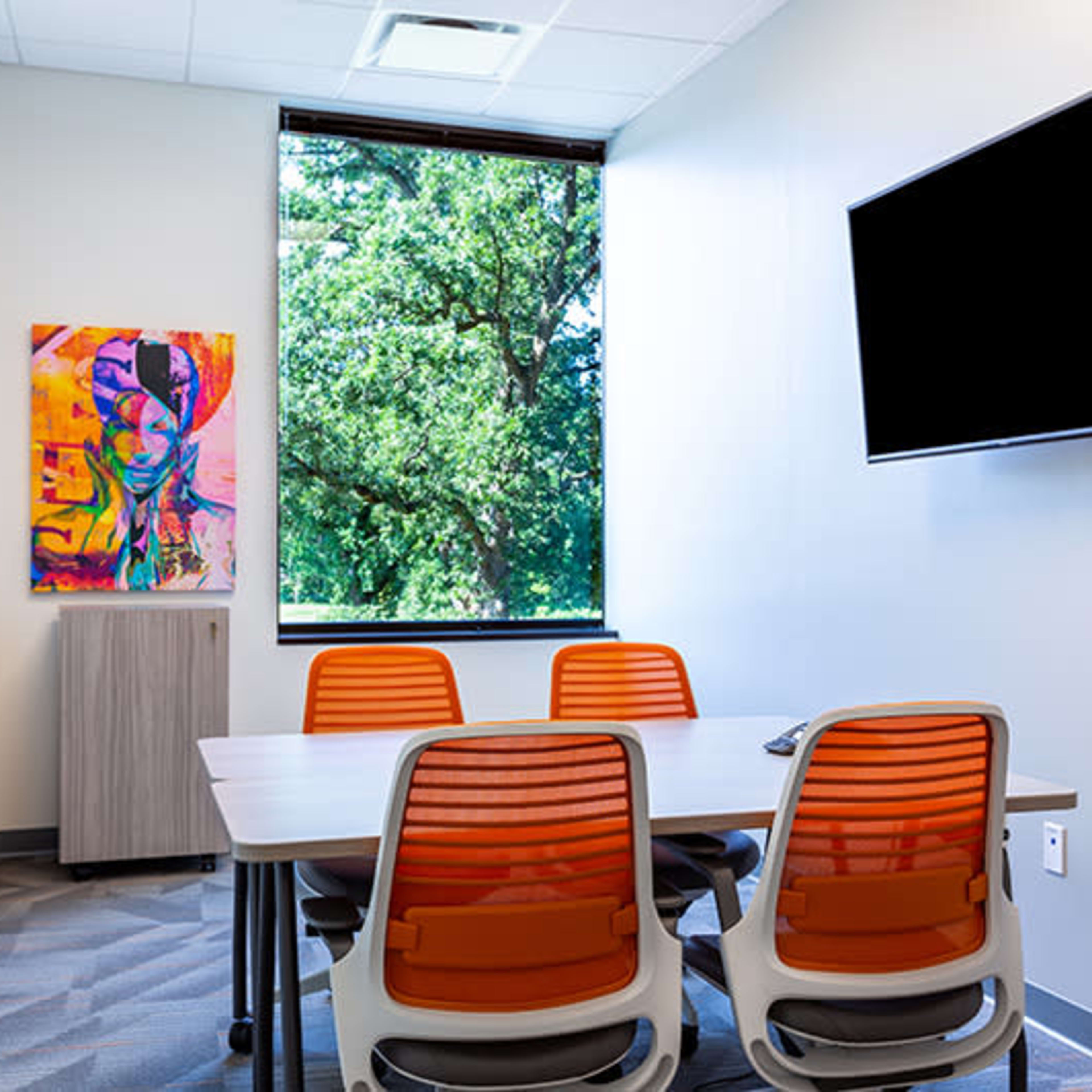 A conference room features a table surrounded by four orange chairs, a wall-mounted television, and a large window overlooking greenery outside.
