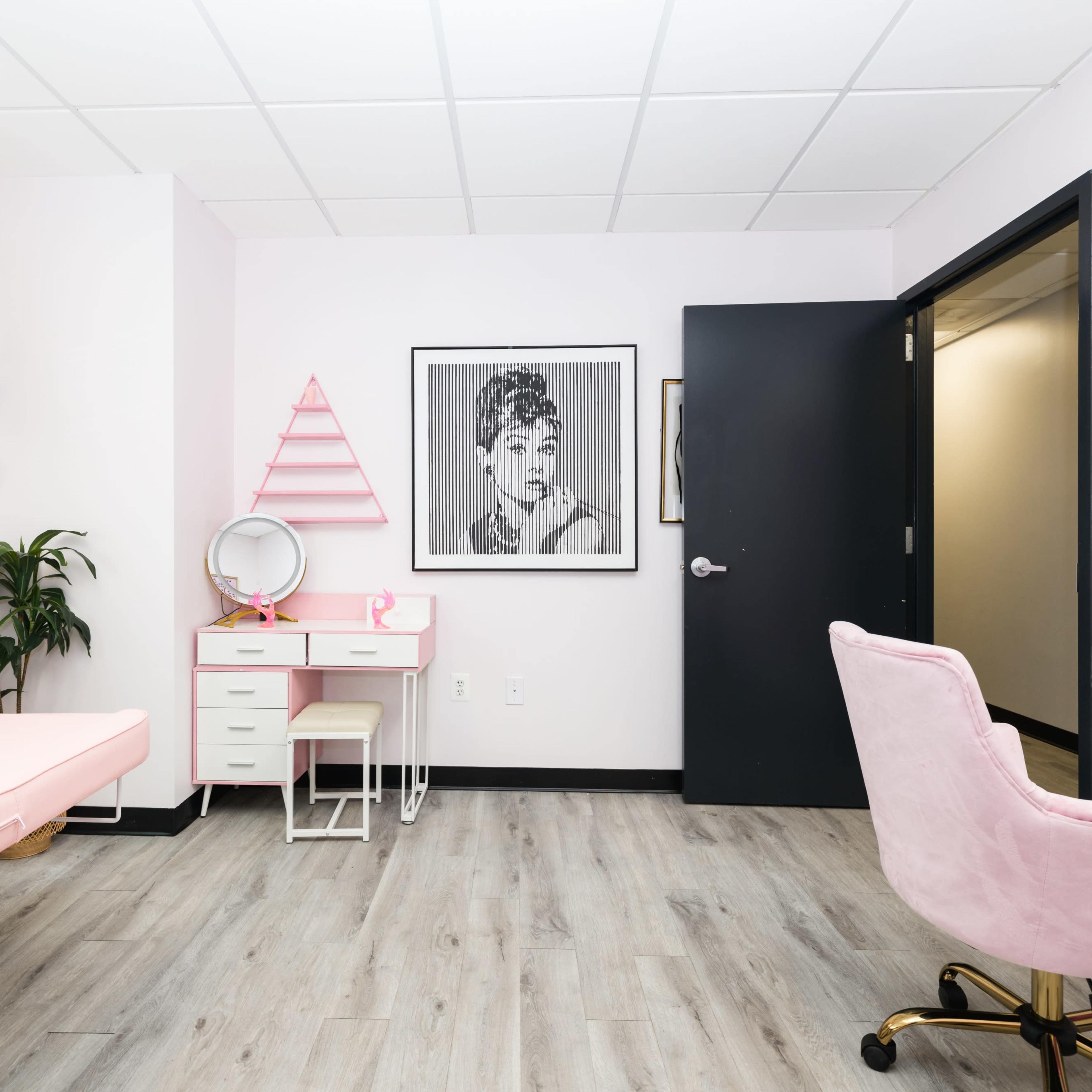 A brightly lit room with pink and white decor, featuring a makeup station, a pink chair, and artwork on the walls.