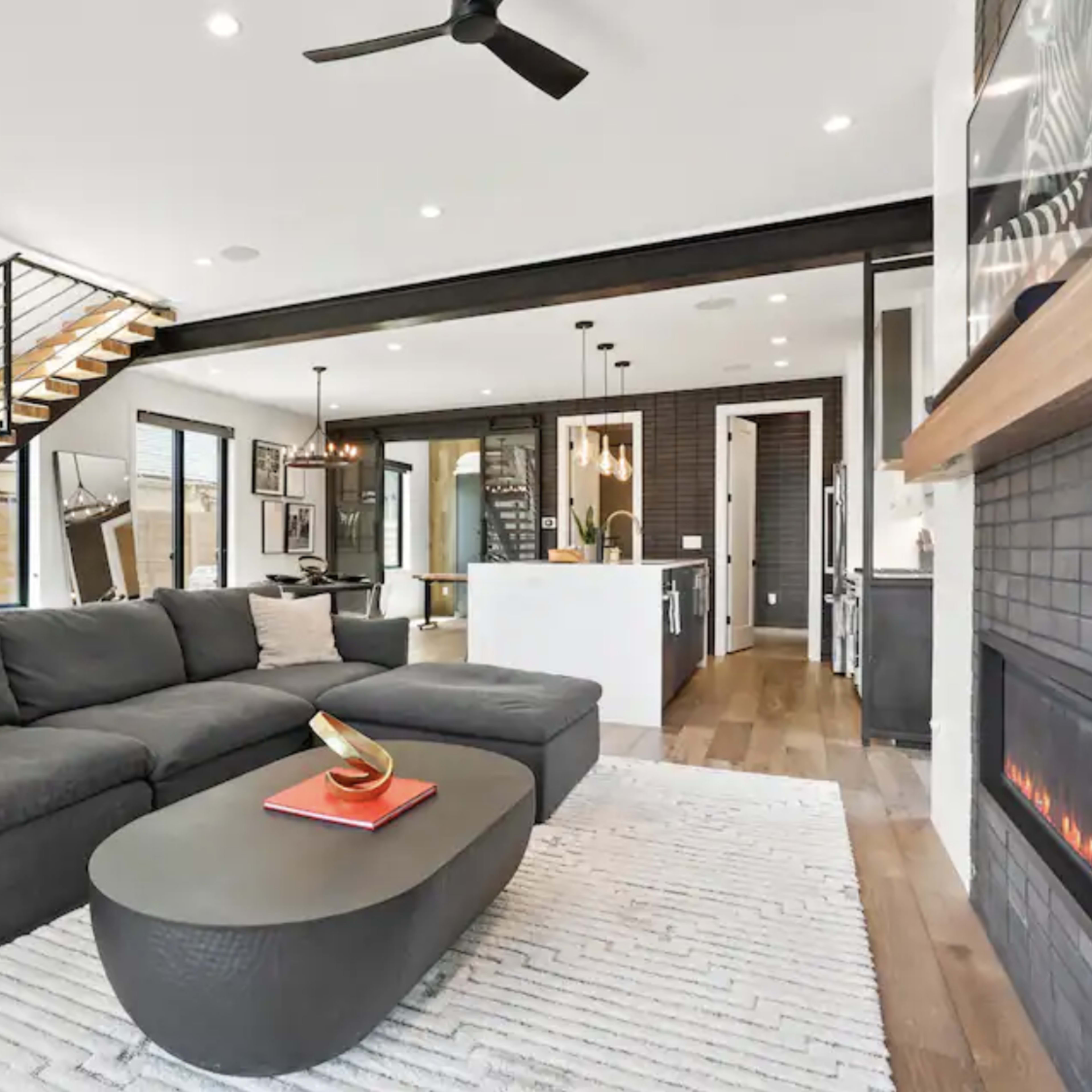 A modern living room features a gray sectional sofa, a round coffee table, a sleek fireplace, and a view of an open kitchen with a staircase leading to the upper level.