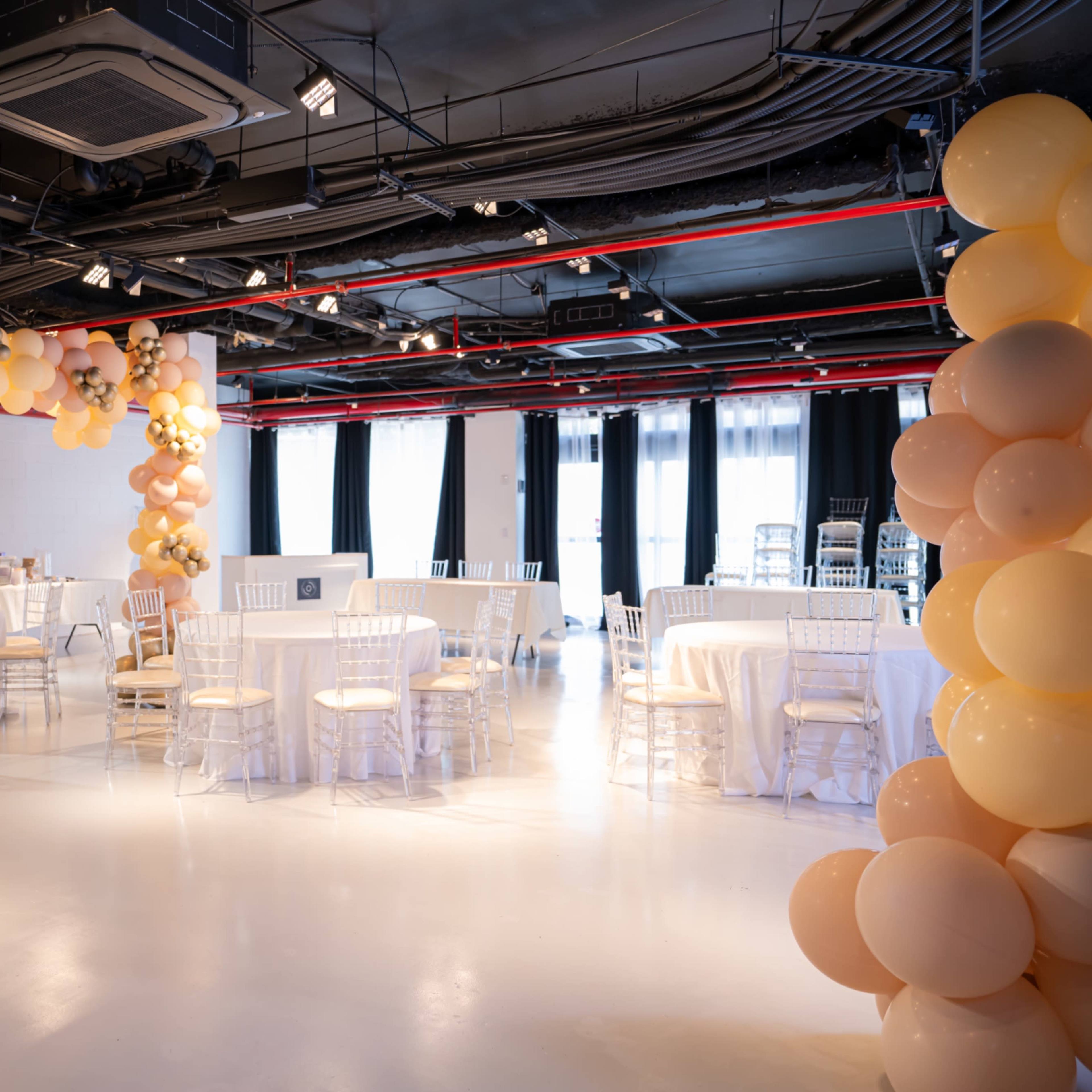 A spacious event room decorated with clusters of balloons and set with circular tables and chairs.