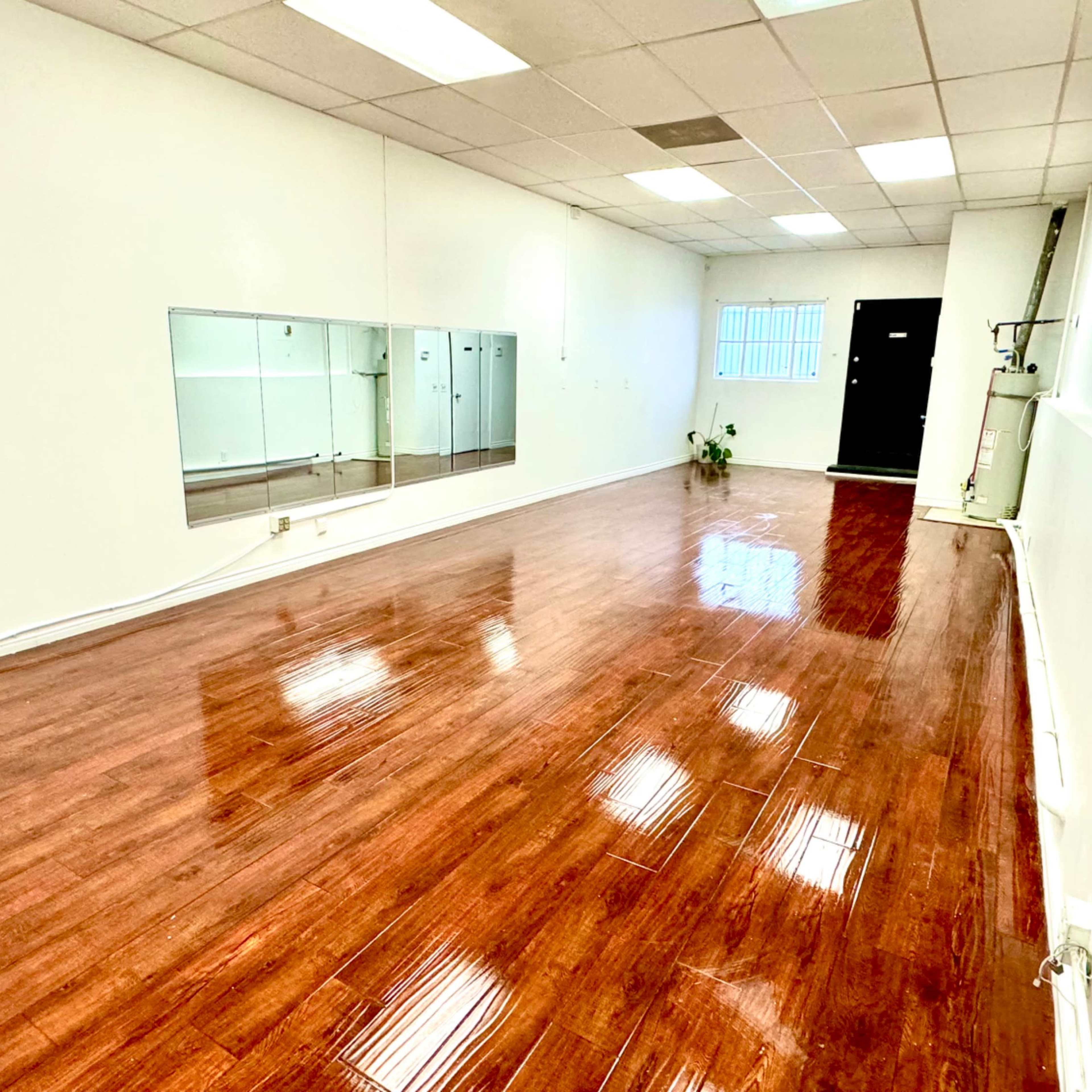 A spacious, well-lit room with polished wooden flooring, mirrors on one wall, and an entrance at the far end.