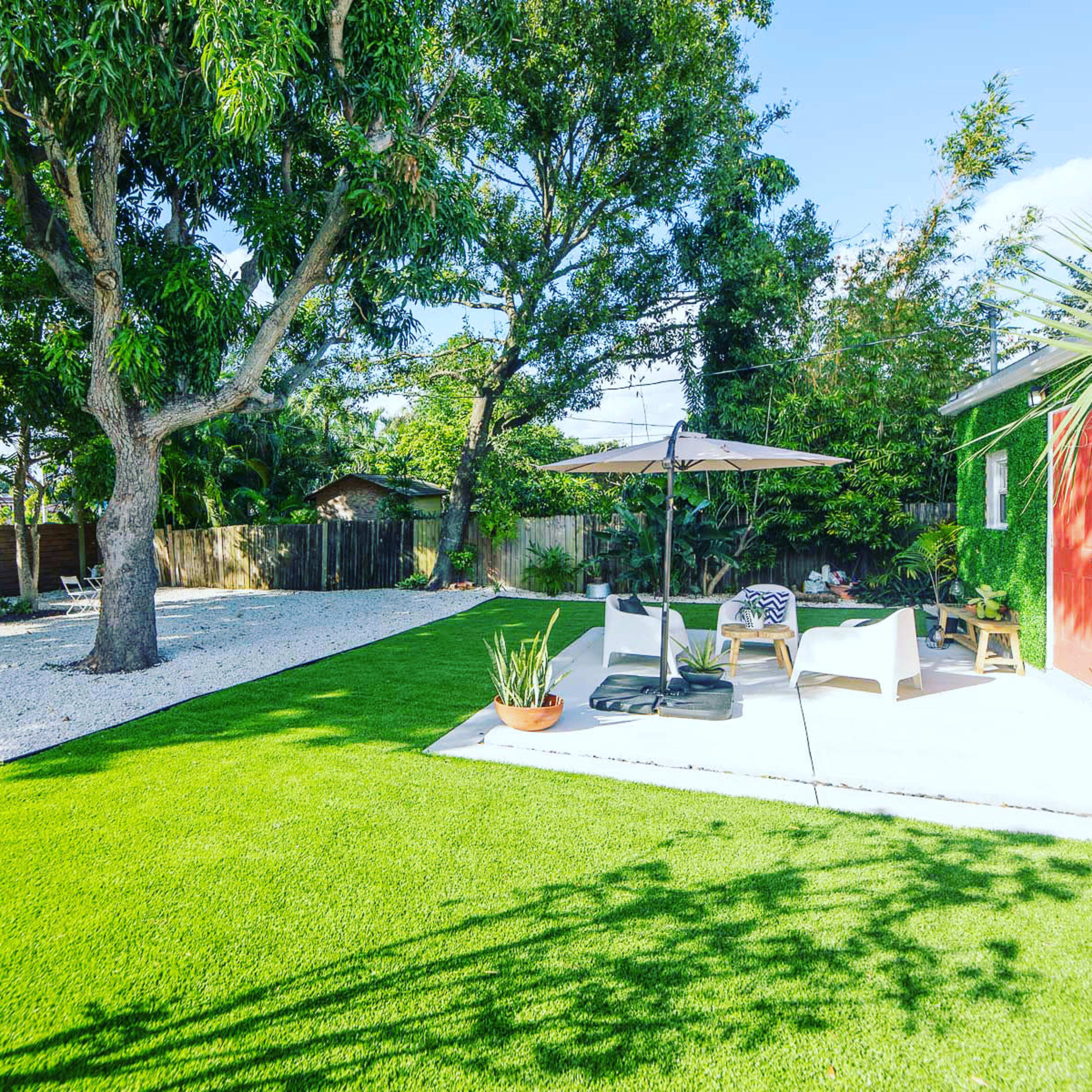 A well-maintained backyard featuring a grassy area, a patio with seating, and trees surrounded by decorative plants.