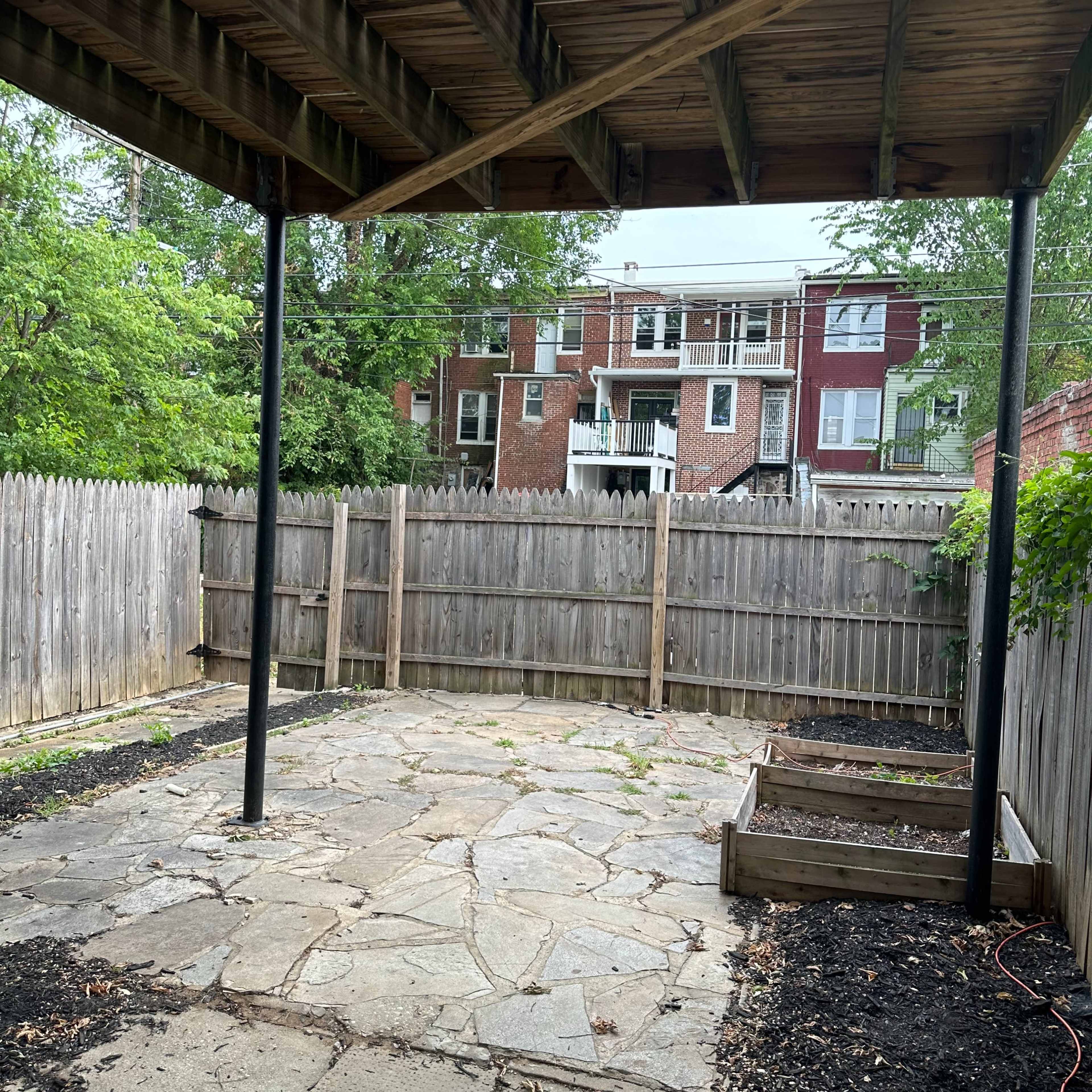 A stone patio area enclosed by a wooden fence with two vertical support beams and a raised garden bed in the corner.