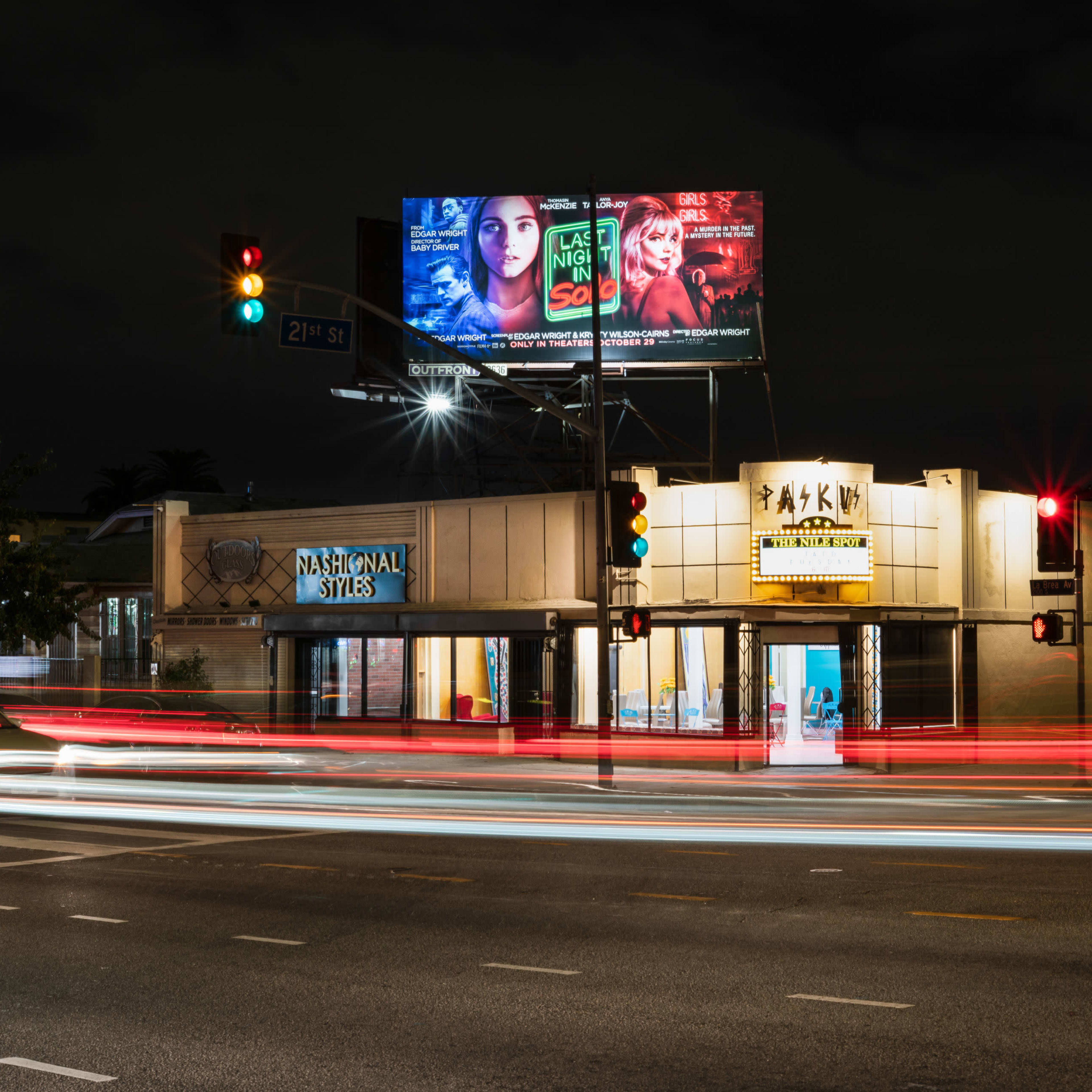 A busy street corner at night features a building with a "National Stores" sign and illuminated billboards above advertising various products.