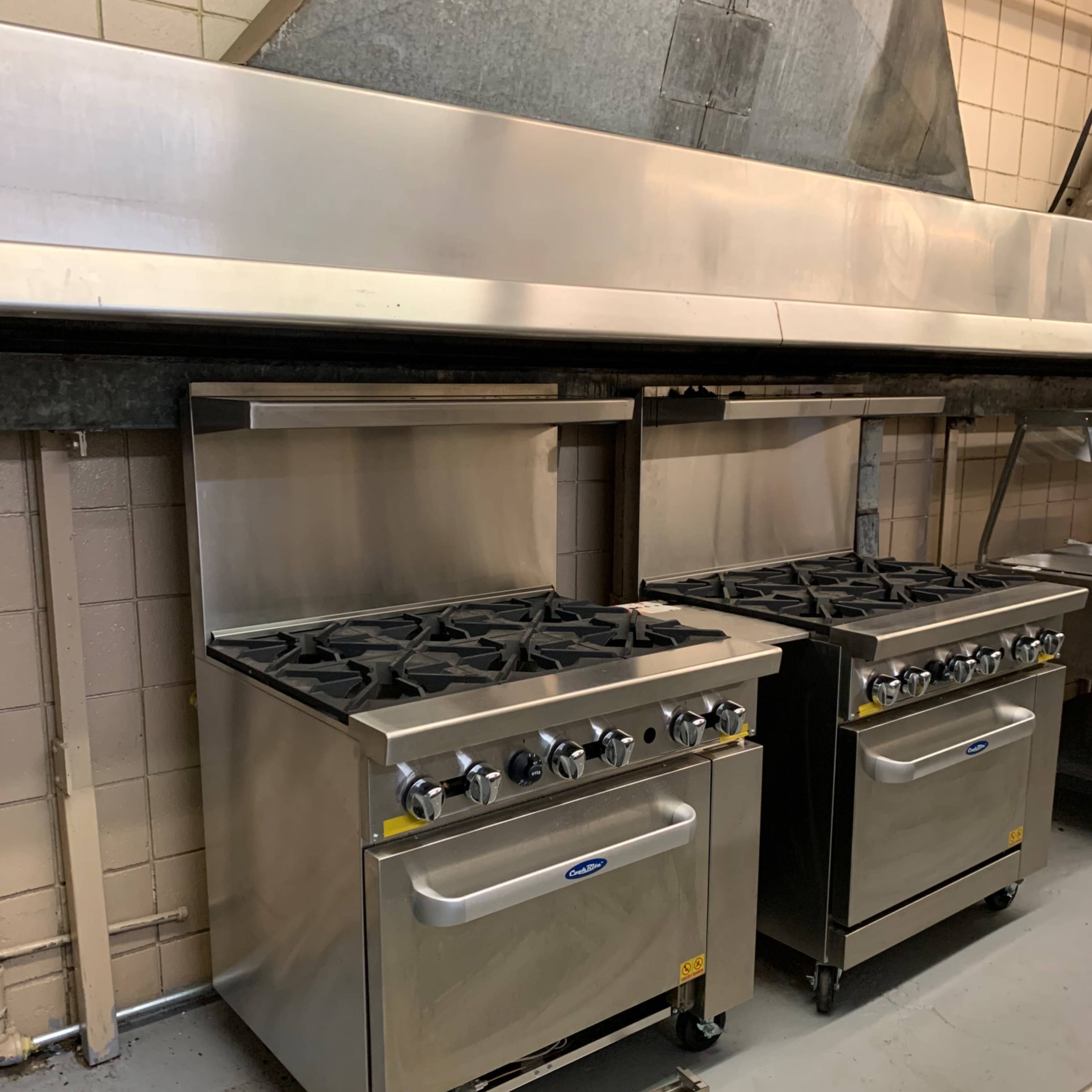 Two stainless steel kitchen ranges with gas burners placed against a tiled wall in a commercial kitchen.