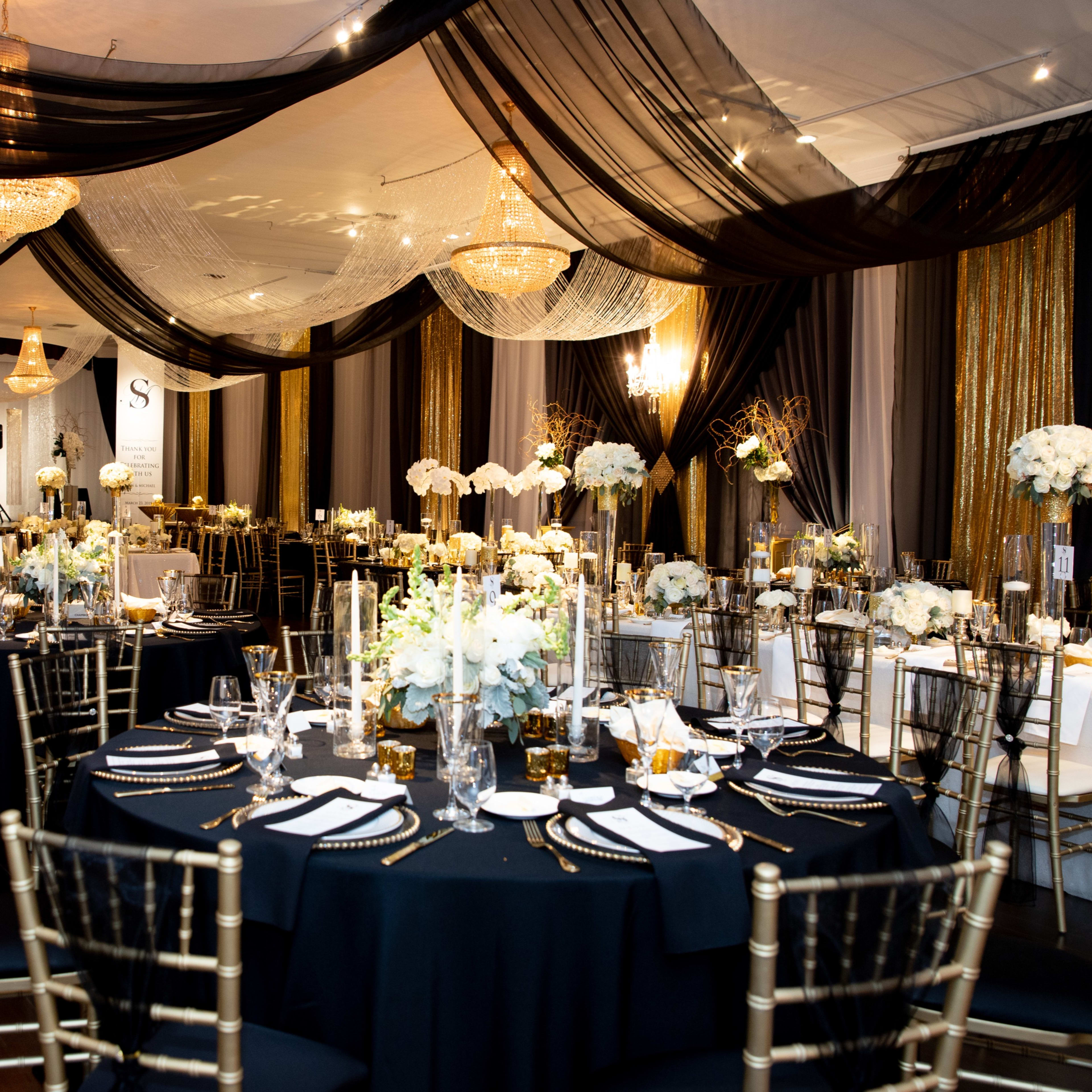 The scene depicts an elegantly decorated event space with round tables set for a formal dinner, adorned with white floral arrangements and gold accents.