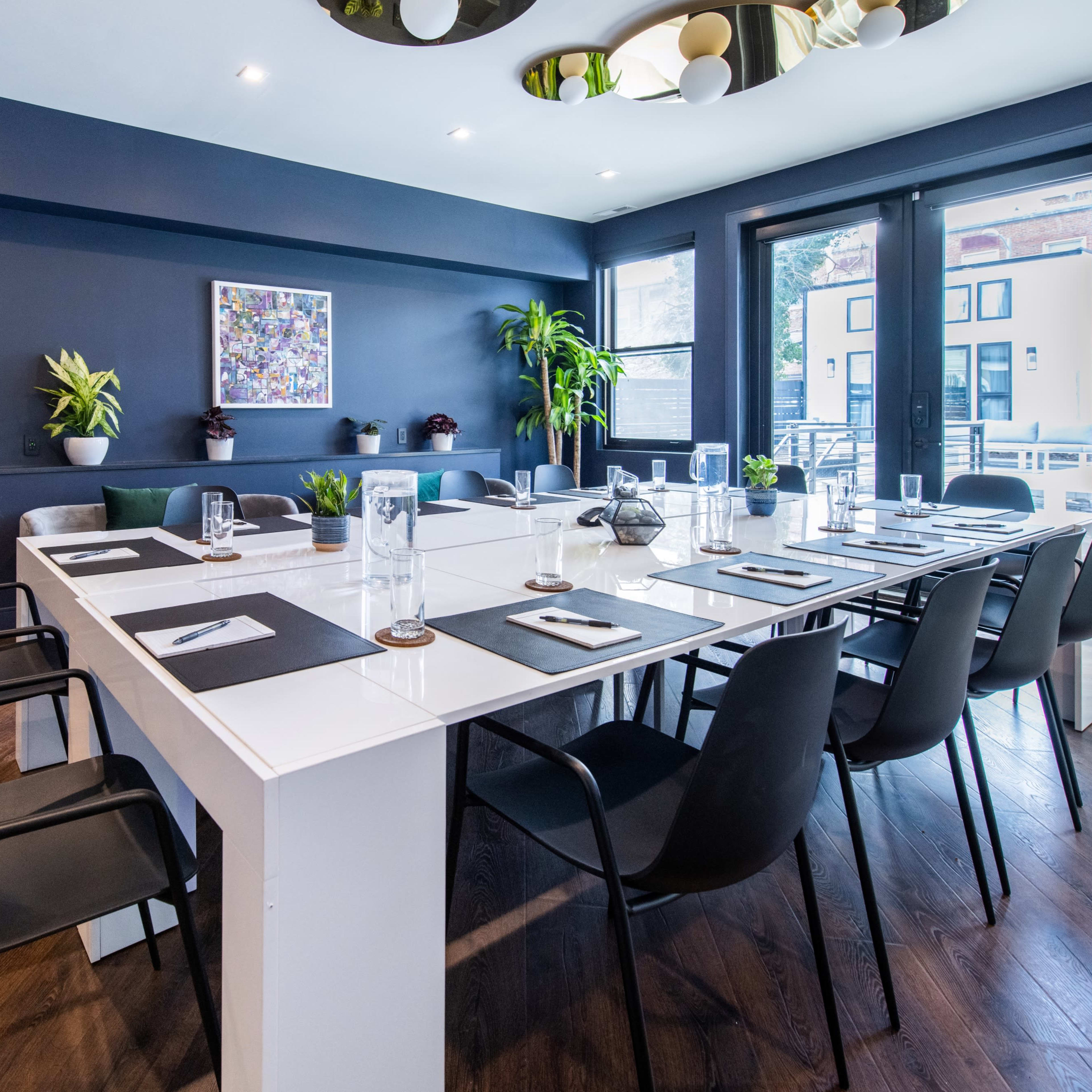 A modern conference room features a large white table set for a meeting, surrounded by black chairs, with plants and artwork along the walls.